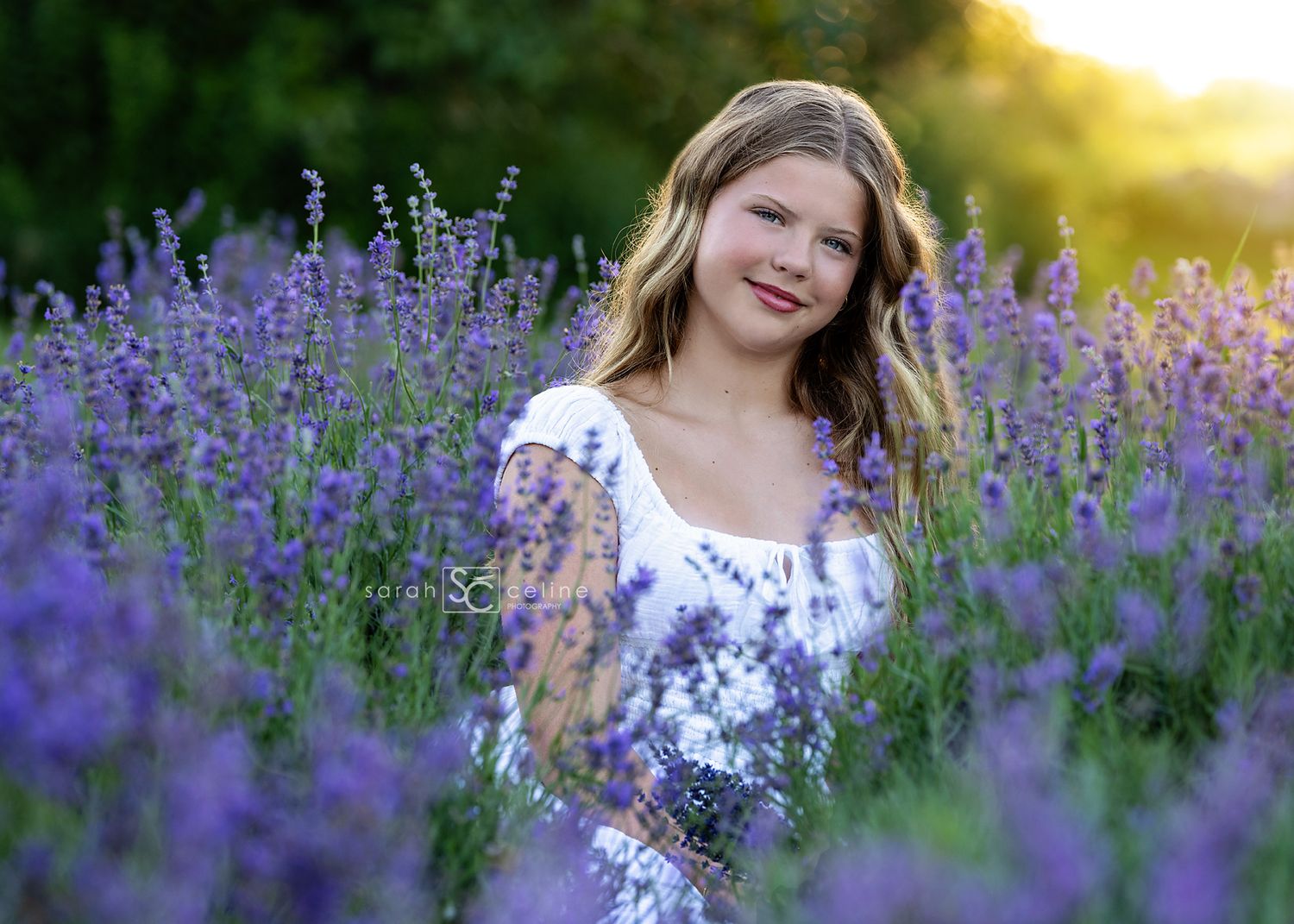 A smiling girl in a white floral dress stands among vibrant purple lavender flowers in a sunlit field.