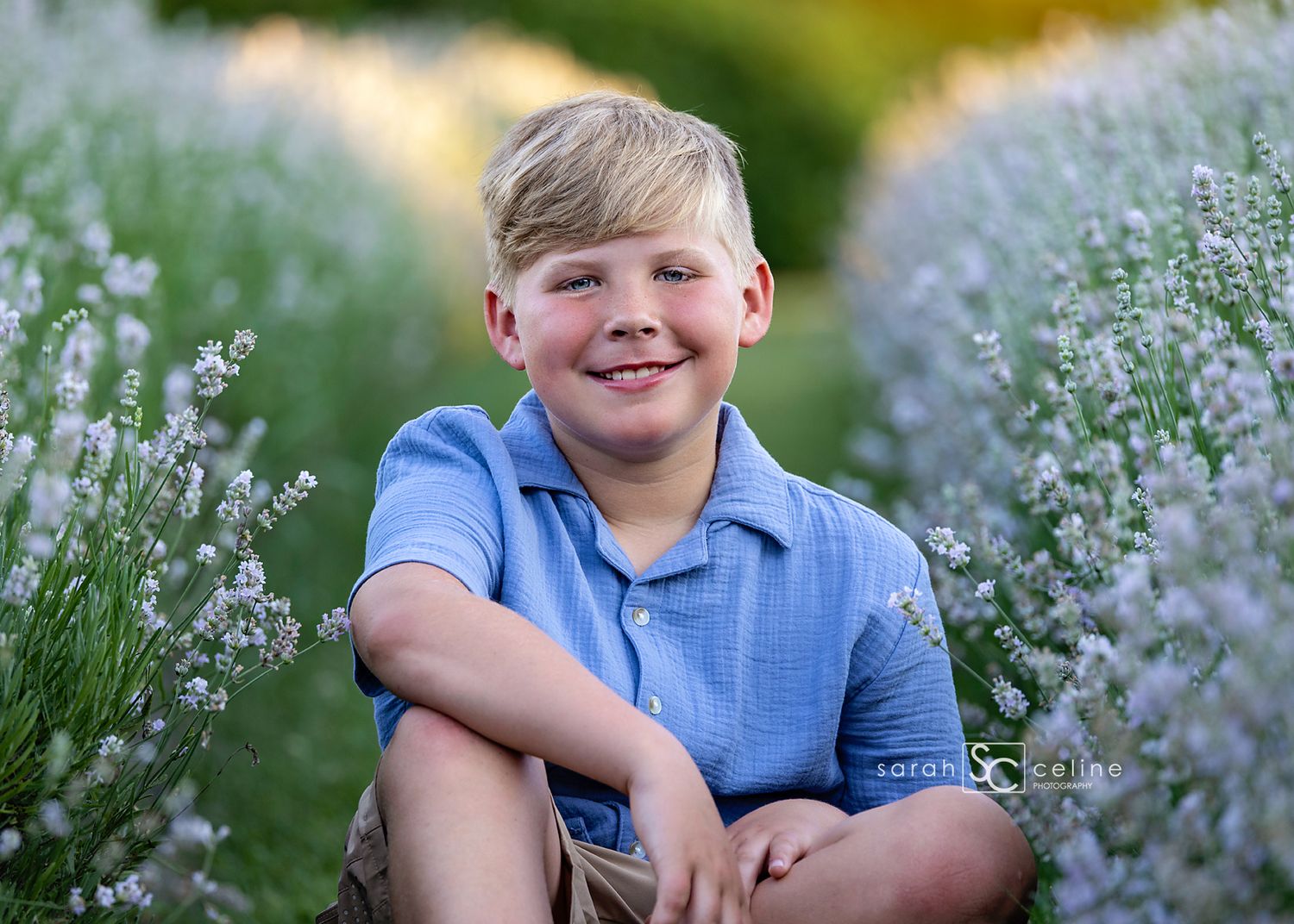 Smiling blond boy in blue polo shirt sitting among purple lavender flowers in a sunny outdoor field.