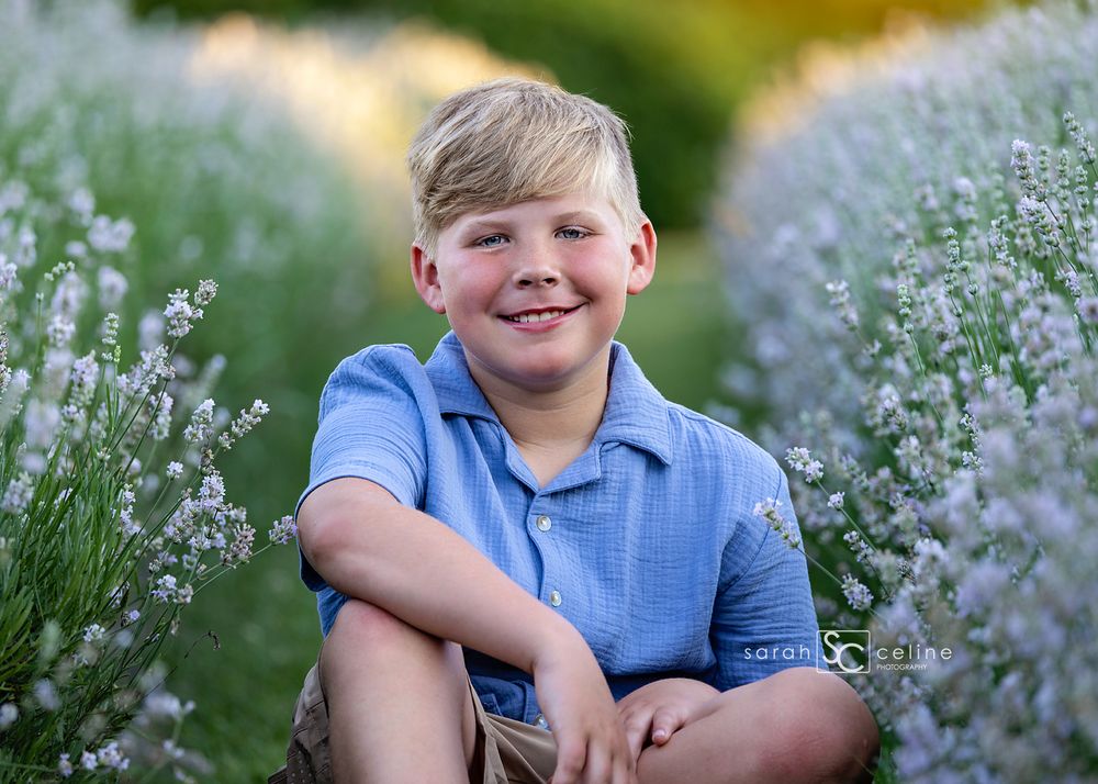 Smiling blond boy in blue polo shirt sitting among purple lavender flowers in a sunny outdoor field.