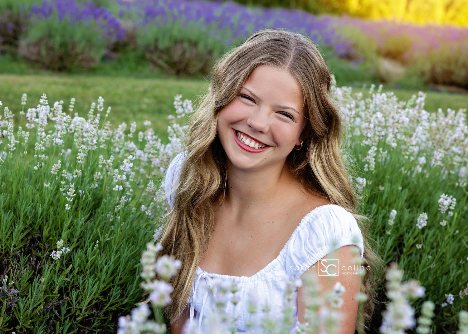 Smiling teenage girl with wavy blonde hair in a white dress posing in a vibrant lavender flower field.