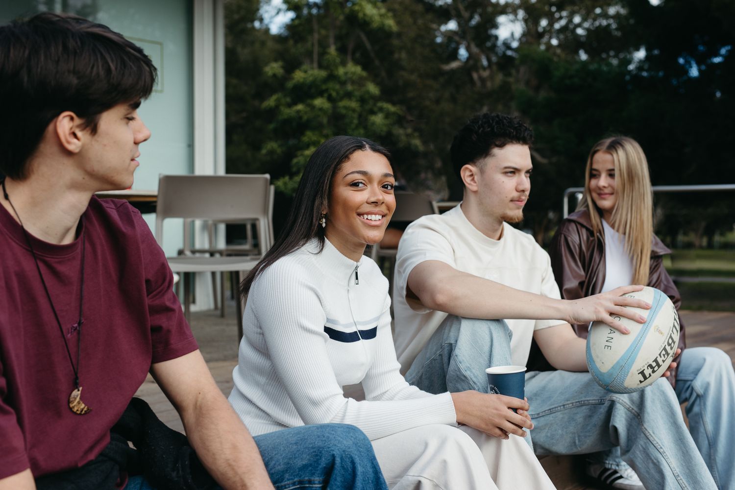 Four diverse young adults sitting outdoors, smiling and socializing, one holding a rugby ball on a sunny day.