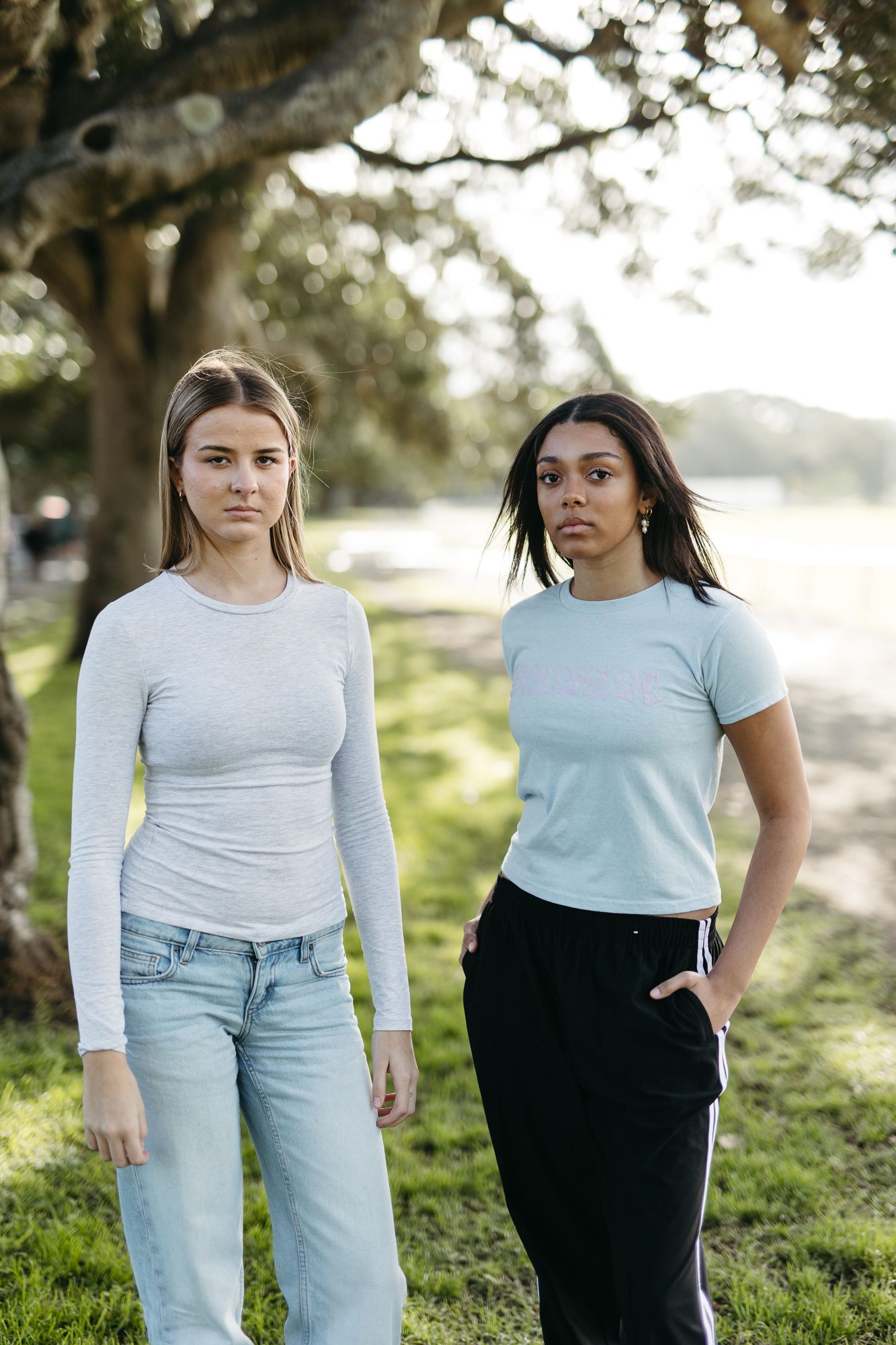 Two young women standing outdoors in a park wearing casual tops and jeans, with trees and greenery in the background.