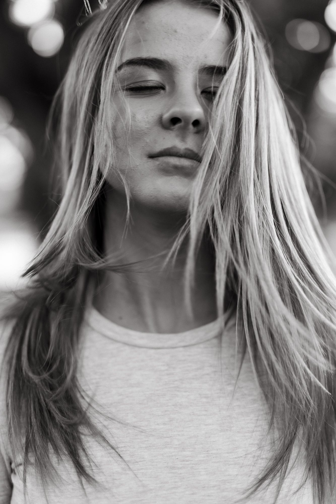 Black and white portrait of a young blonde woman with eyes closed, wearing a white t-shirt, hair blowing gently.