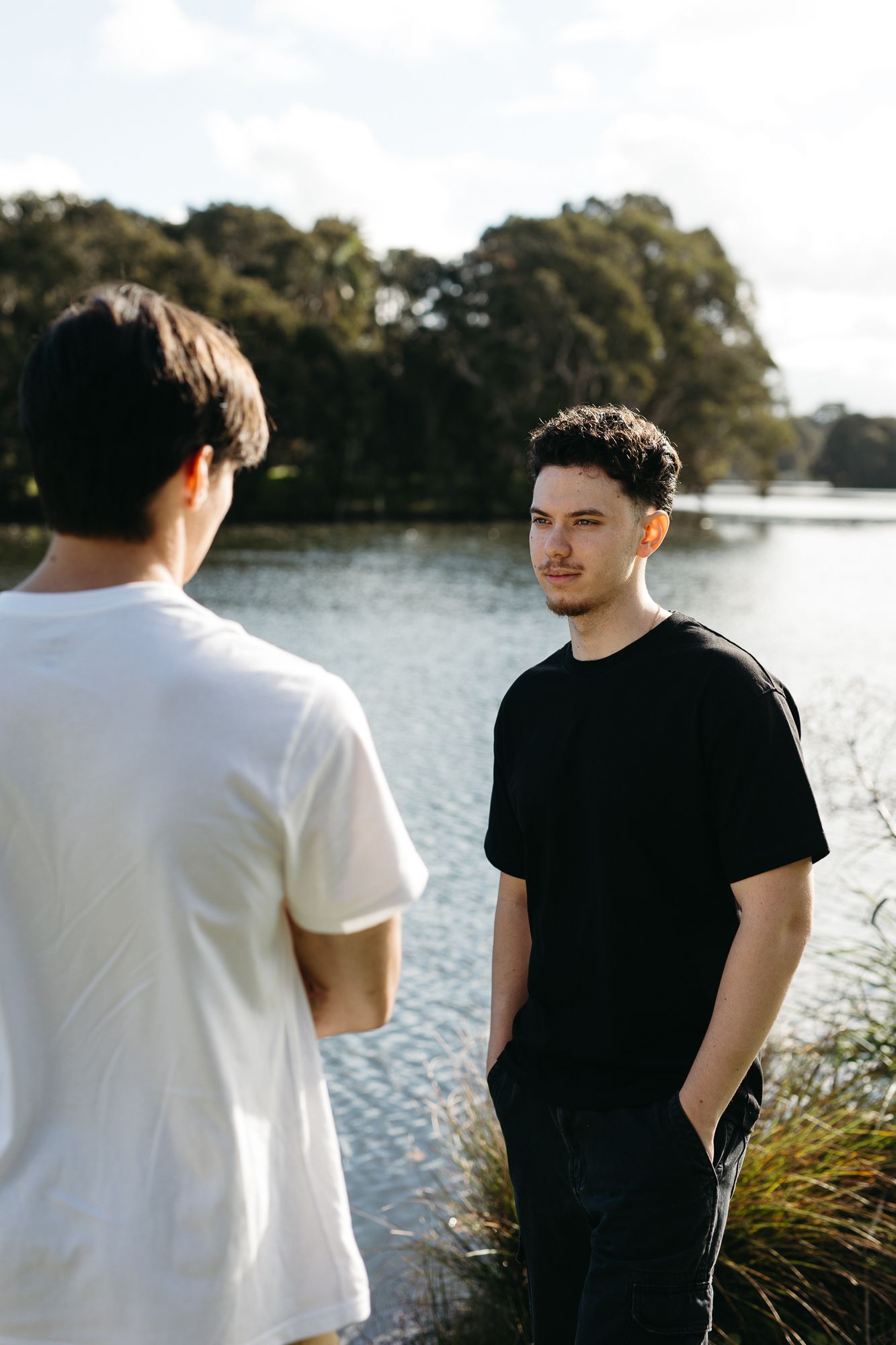 Two young men in casual t-shirts conversing near a scenic lake surrounded by lush green trees on a sunny day.