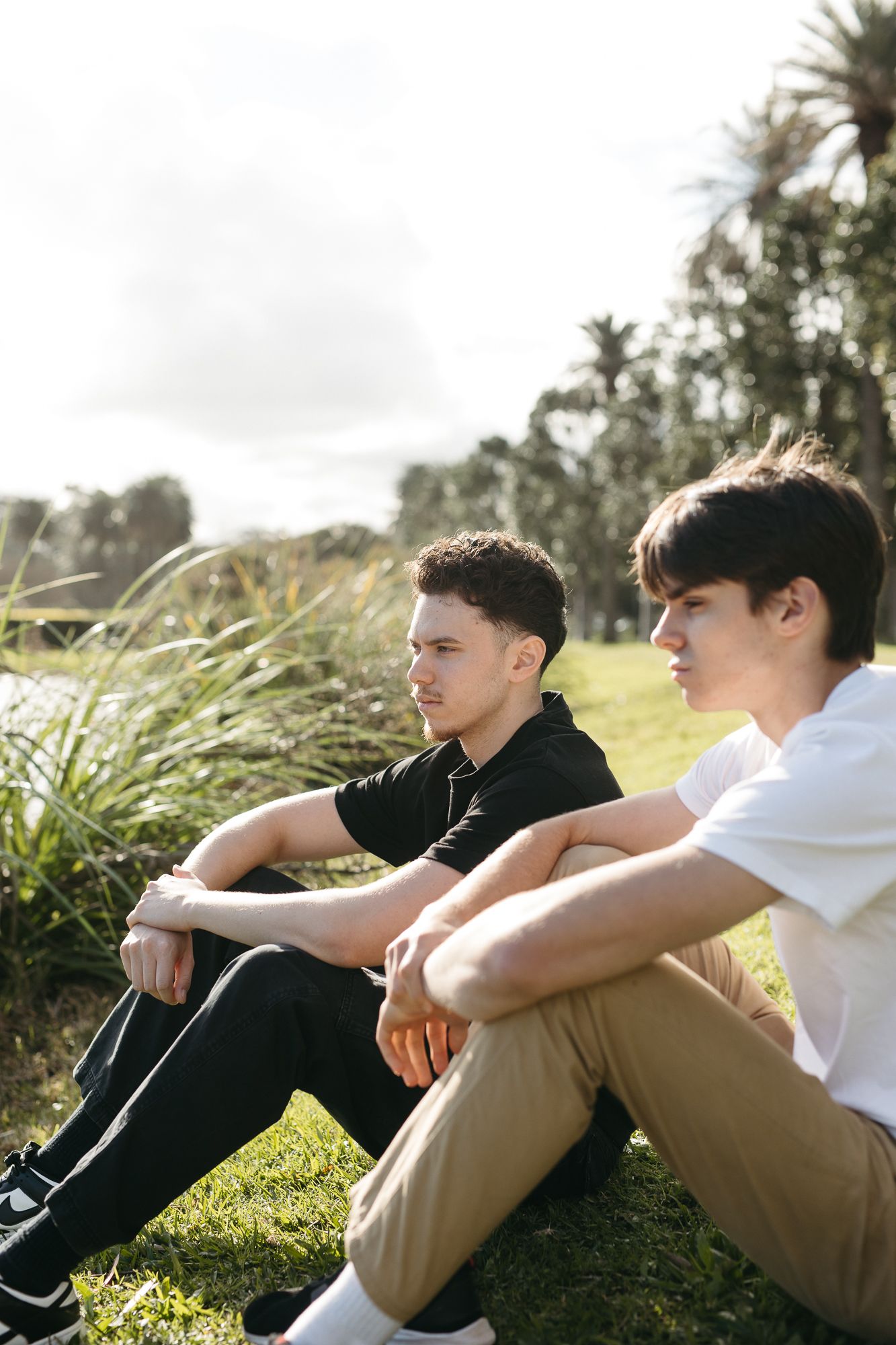 Two young men sitting on grass in a park, relaxing outdoors on a sunny day with trees in the background.