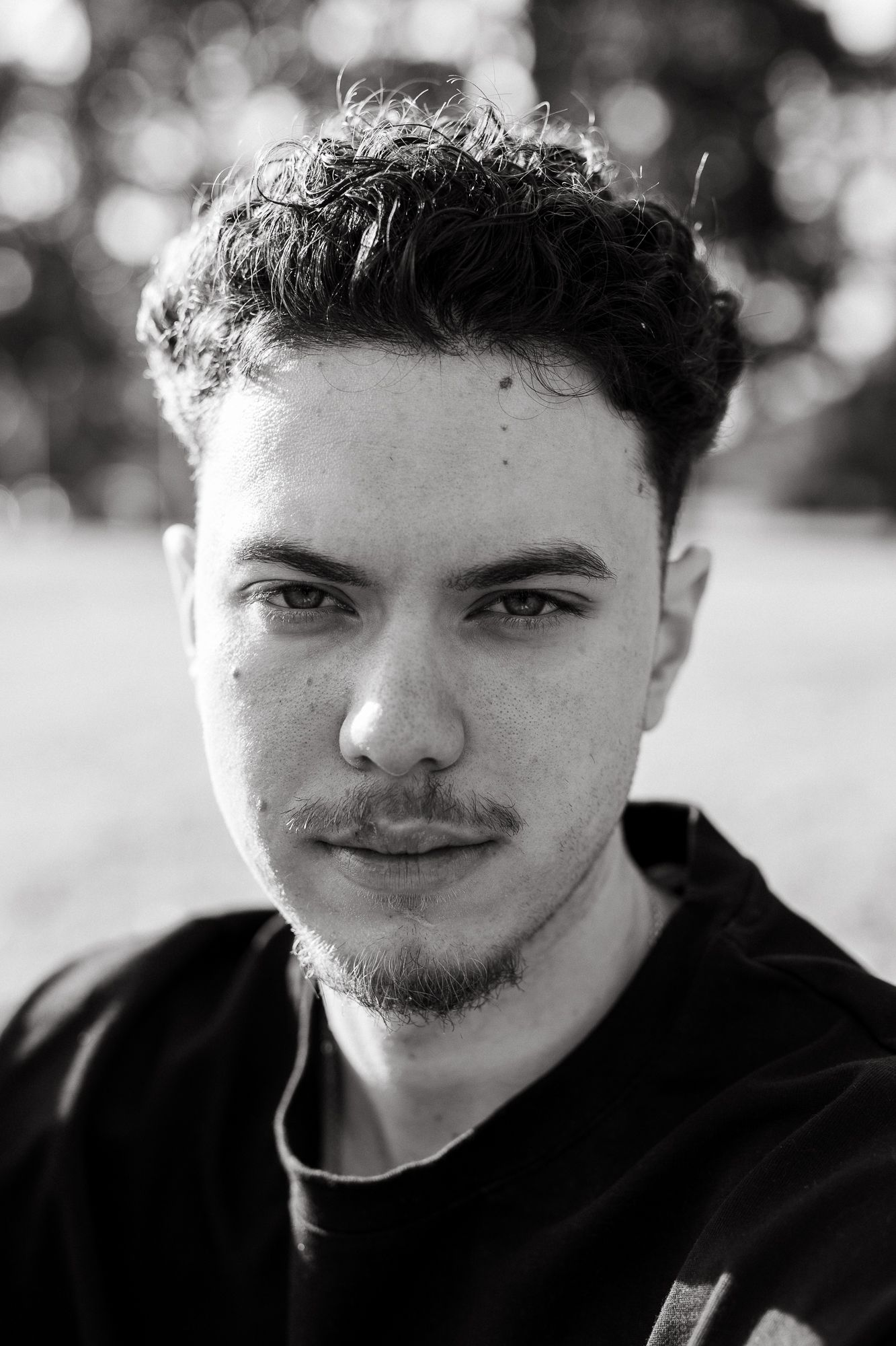 Black and white portrait of a young man with curly hair and light stubble, gazing confidently at the camera outdoors.