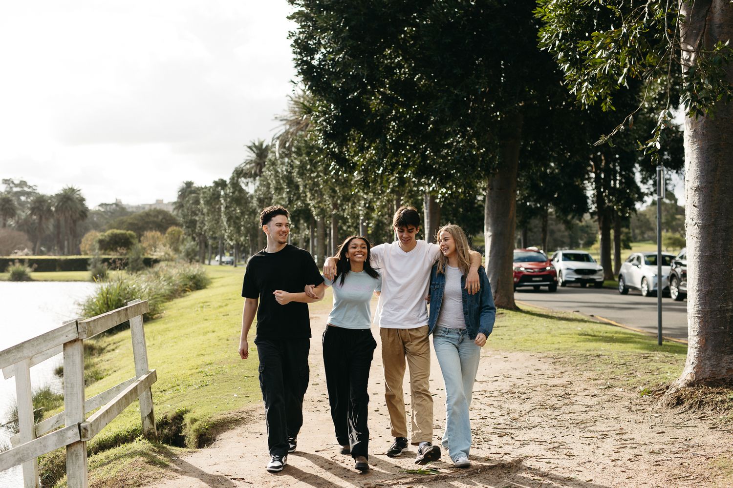 Four friends walking together on a sunny park path lined with trees near a lake.