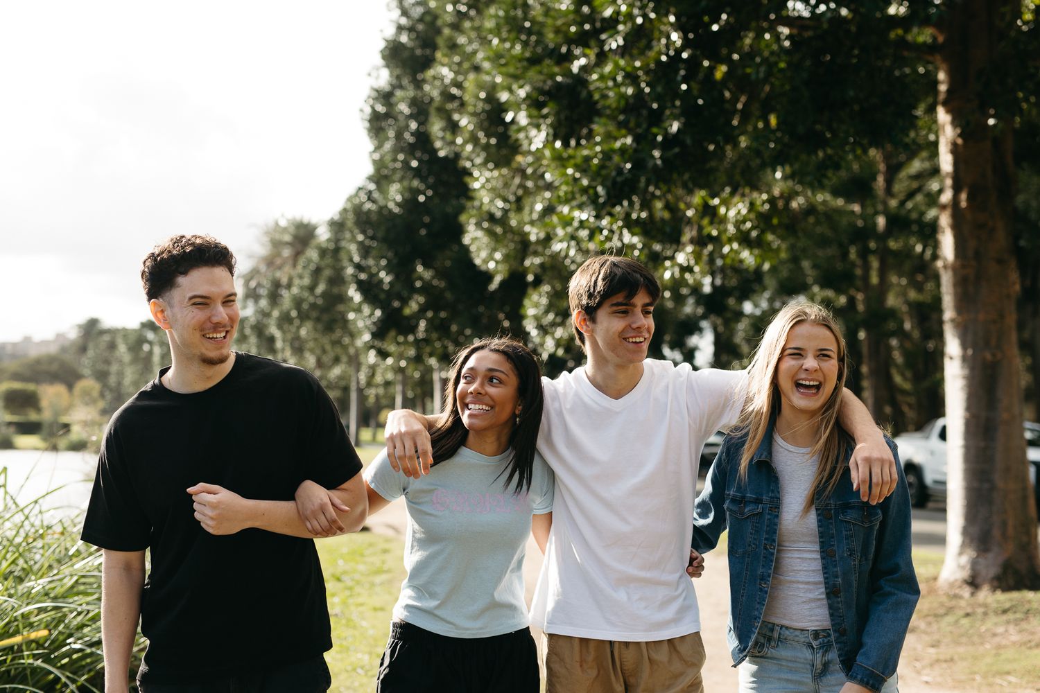 Four happy teenagers walking together outdoors, smiling and embracing on a tree-lined path on a sunny day.