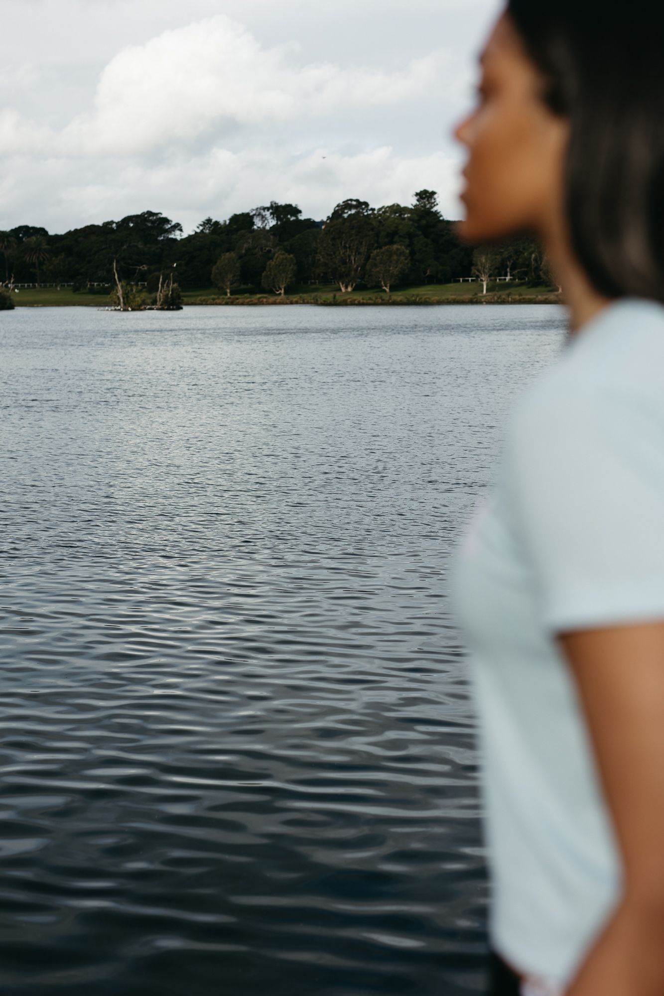 Woman in white t-shirt standing by a calm lake with trees and cloudy sky in the background.