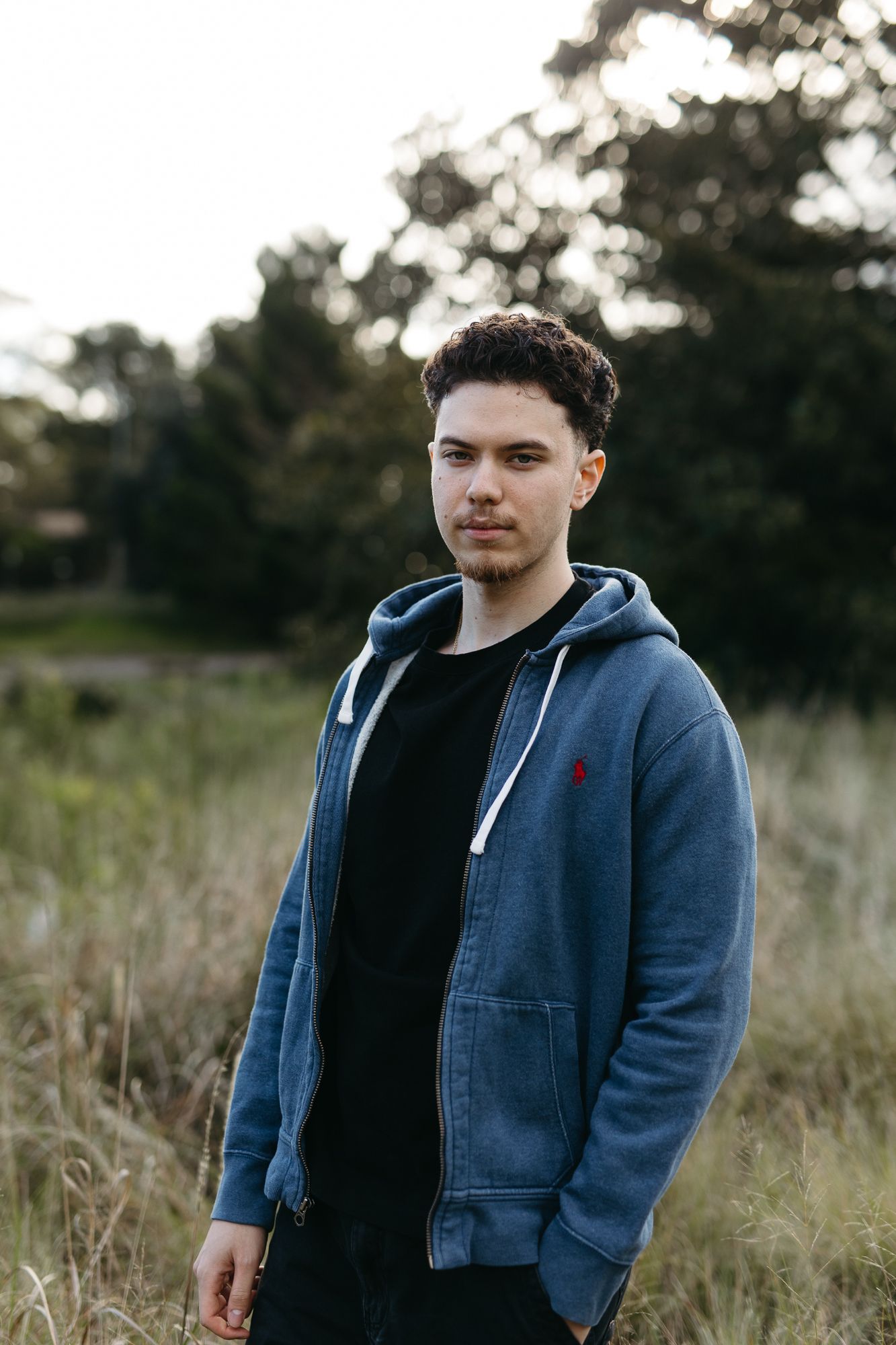 Young man in blue zip-up hoodie standing in tall grass with trees in background.