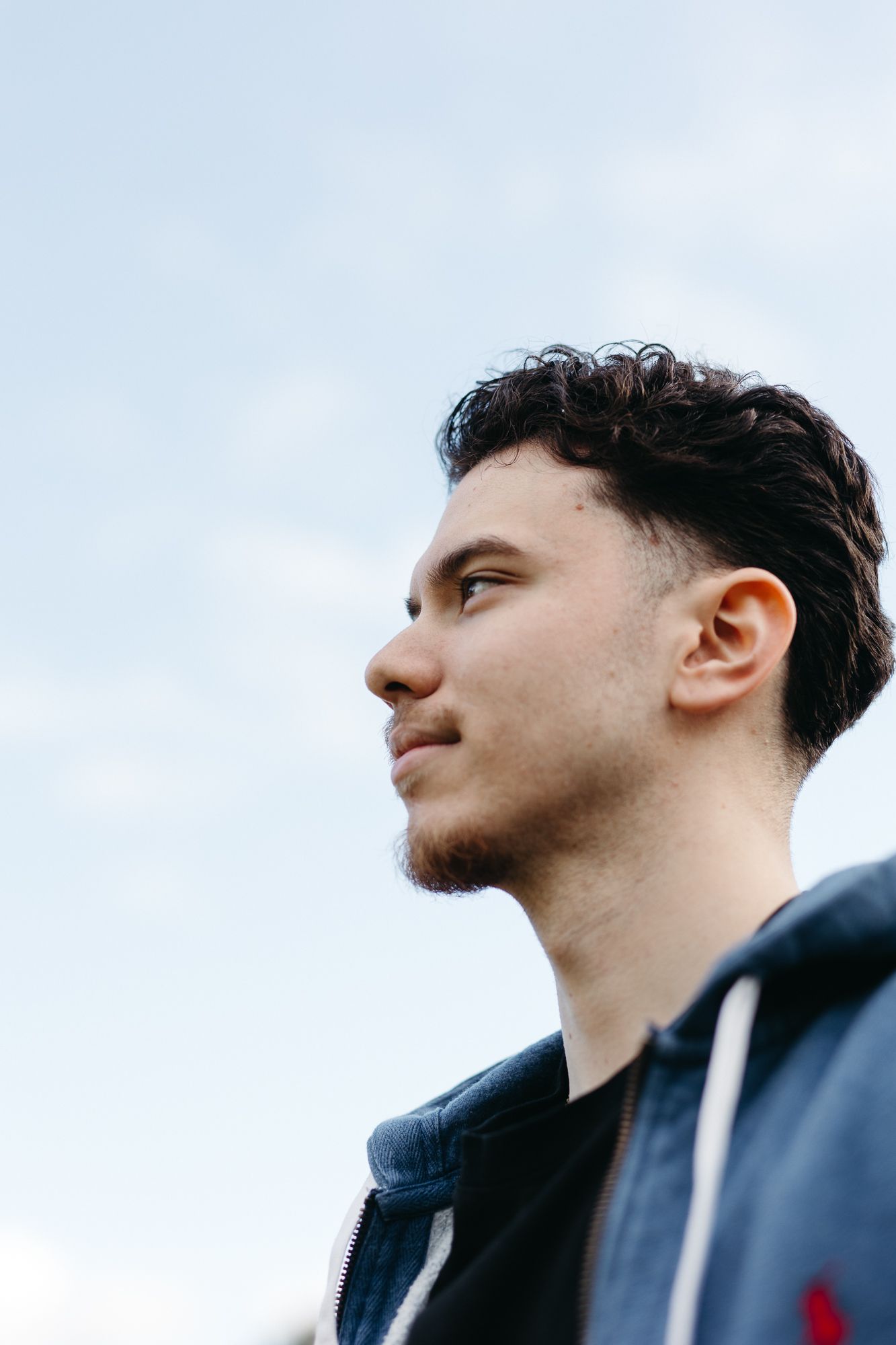 Young man with curly hair and beard gazing thoughtfully against a cloudy sky, wearing a blue jacket.