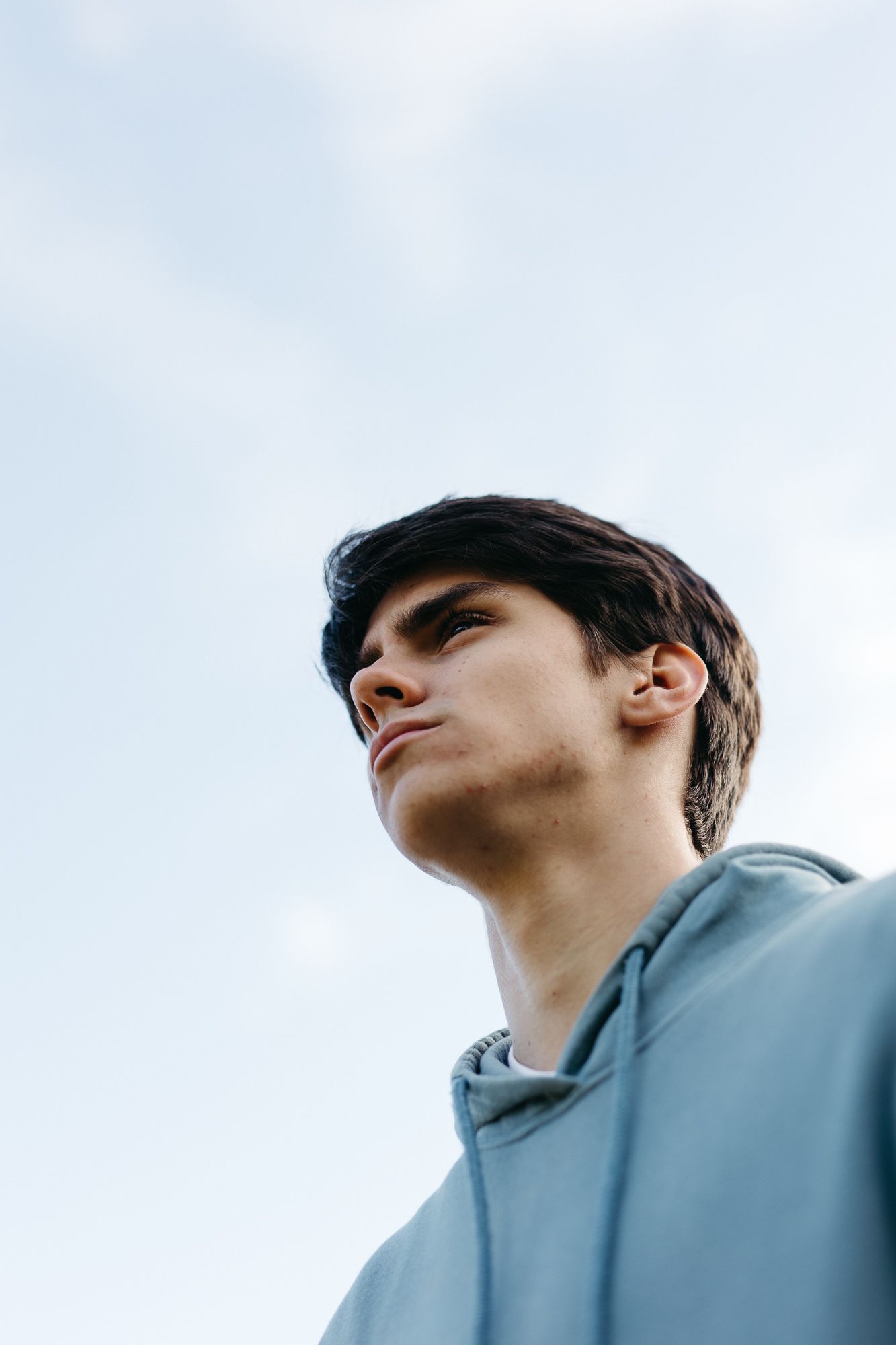 Young man in a light blue hoodie gazing upward against a cloudy sky, shot from a low angle perspective.