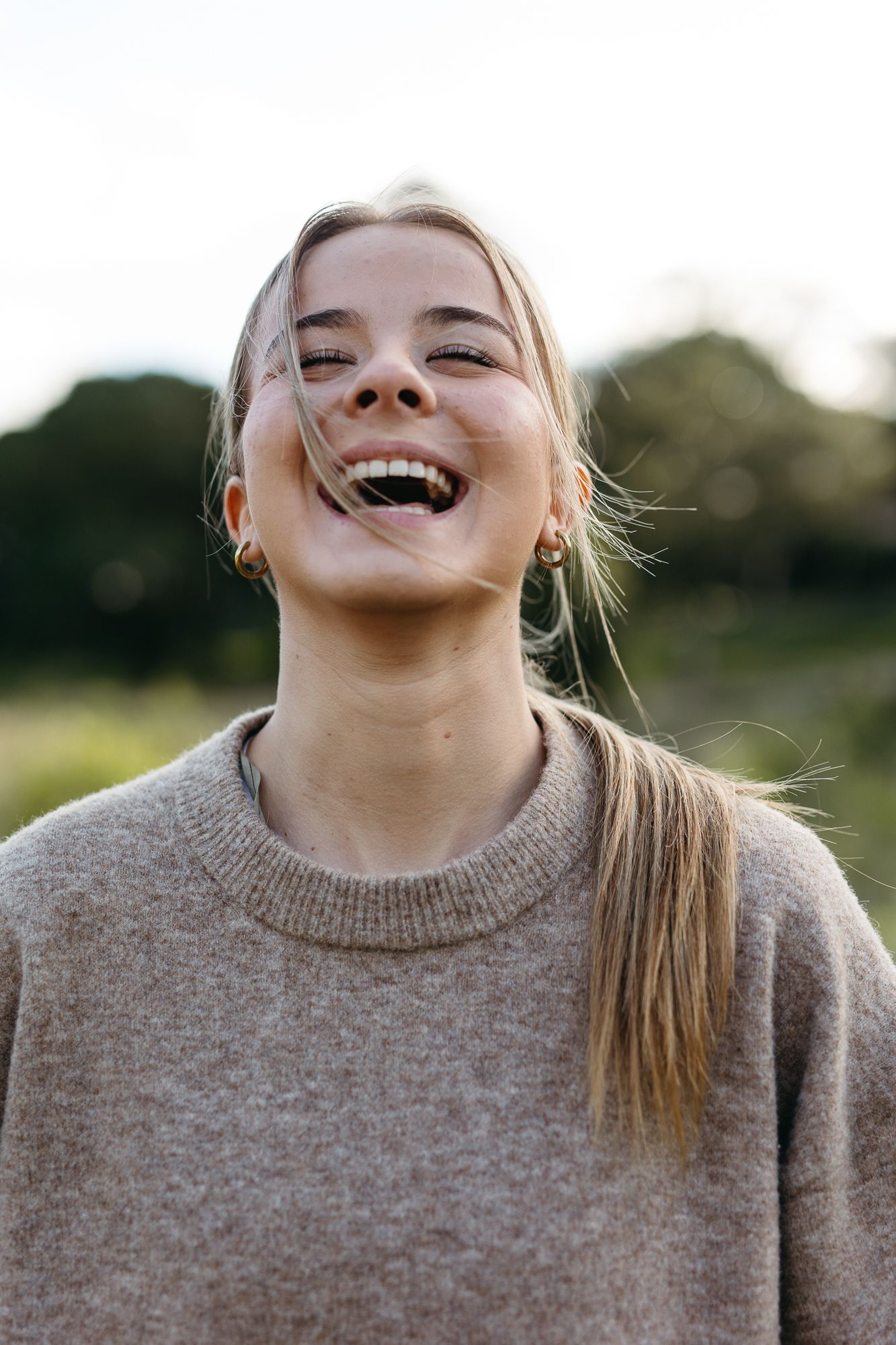 Young blonde woman laughing outdoors in a beige knit sweater, with green trees in the background.