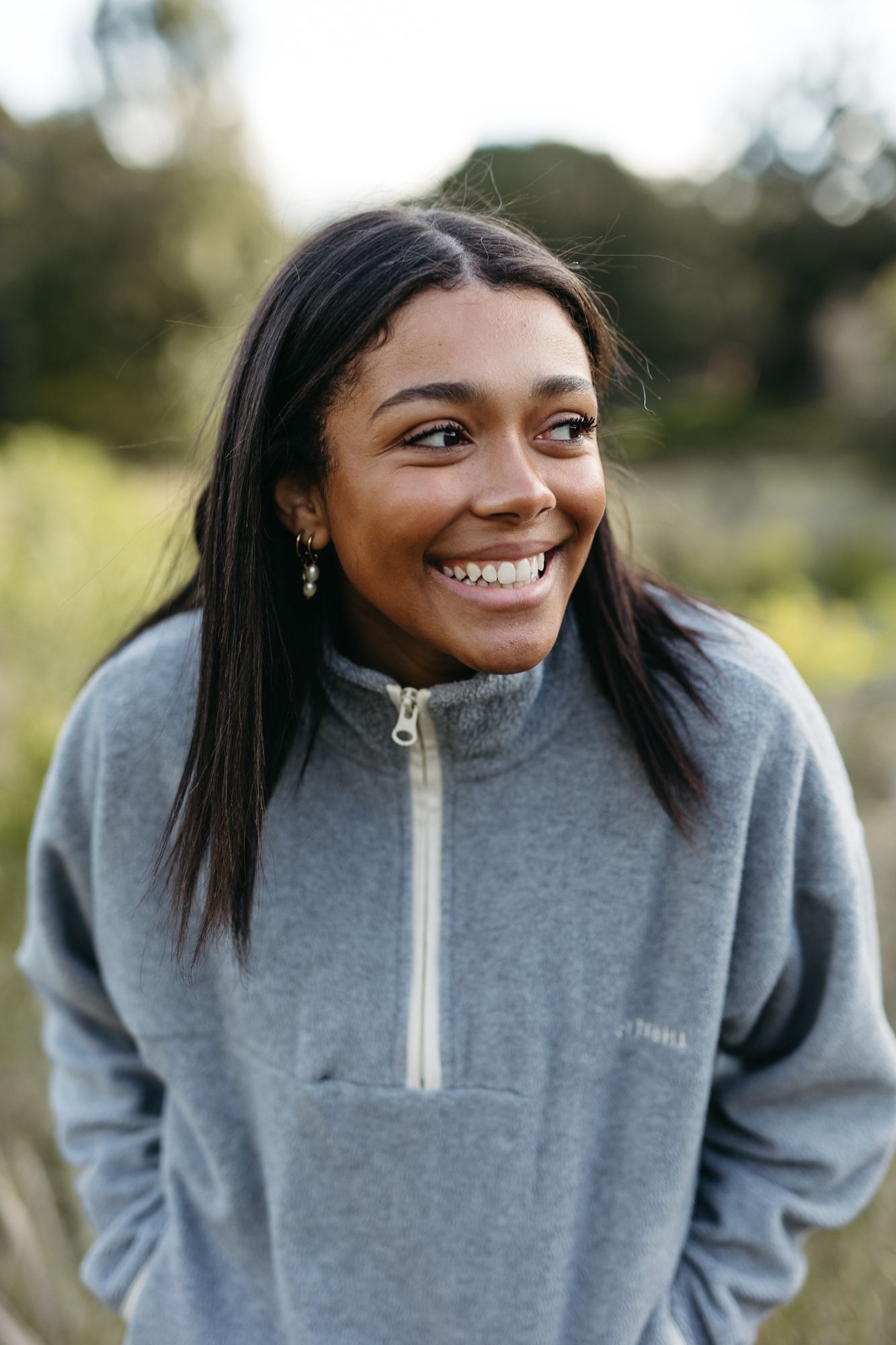 Young woman smiling outdoors in a gray quarter-zip fleece pullover, with long dark hair and gold earrings.