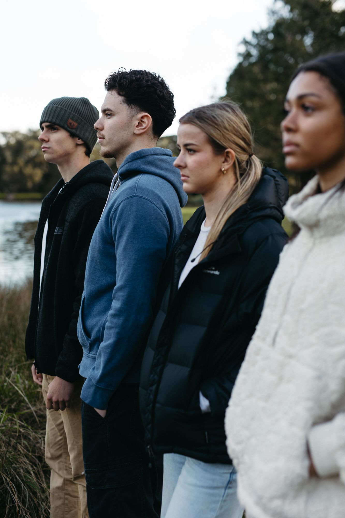 Four young adults standing outdoors near a lake, wearing casual hoodies and jackets, looking serious.