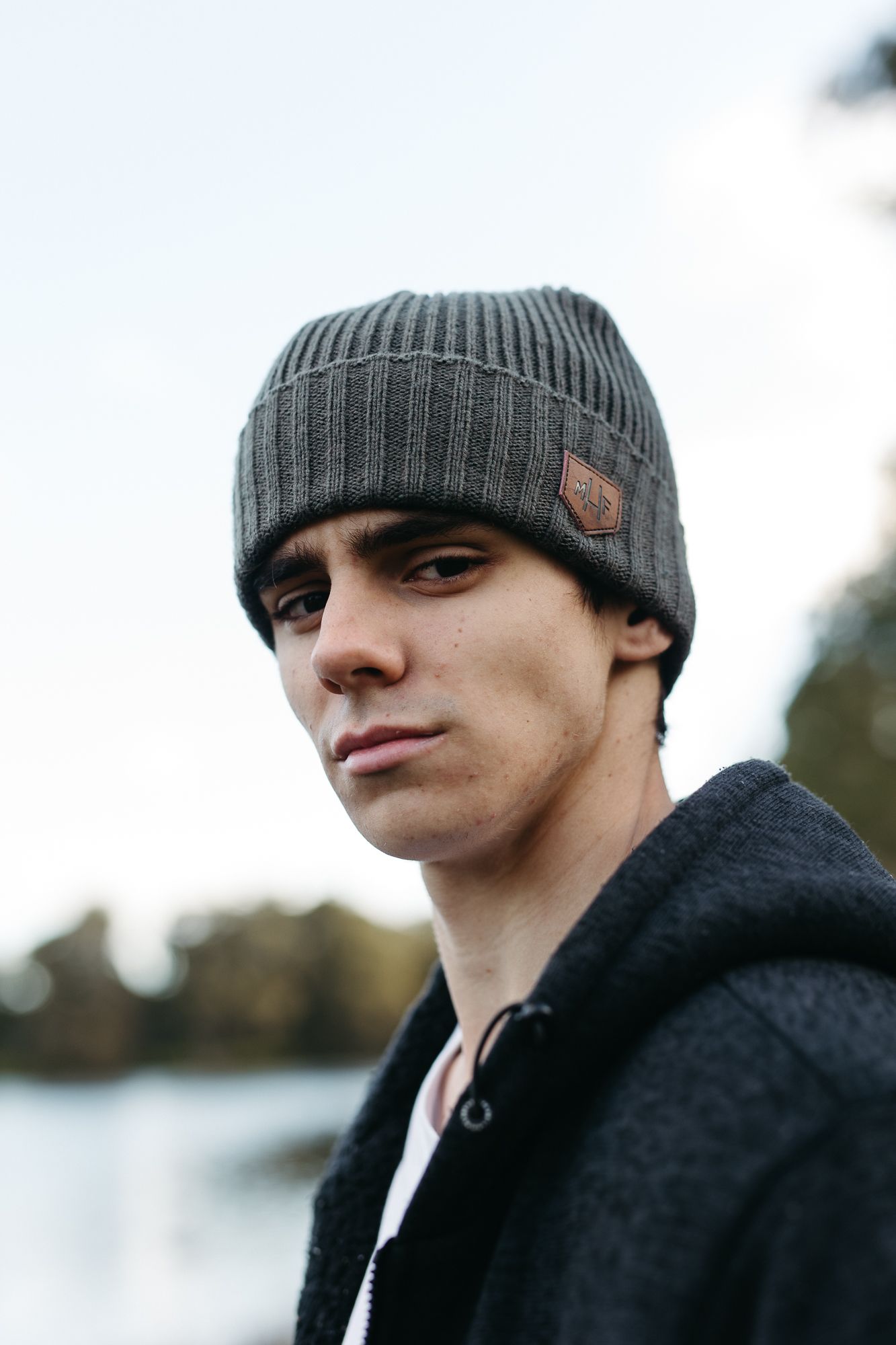 Young man wearing a gray ribbed beanie hat and black coat outdoors near a lake.
