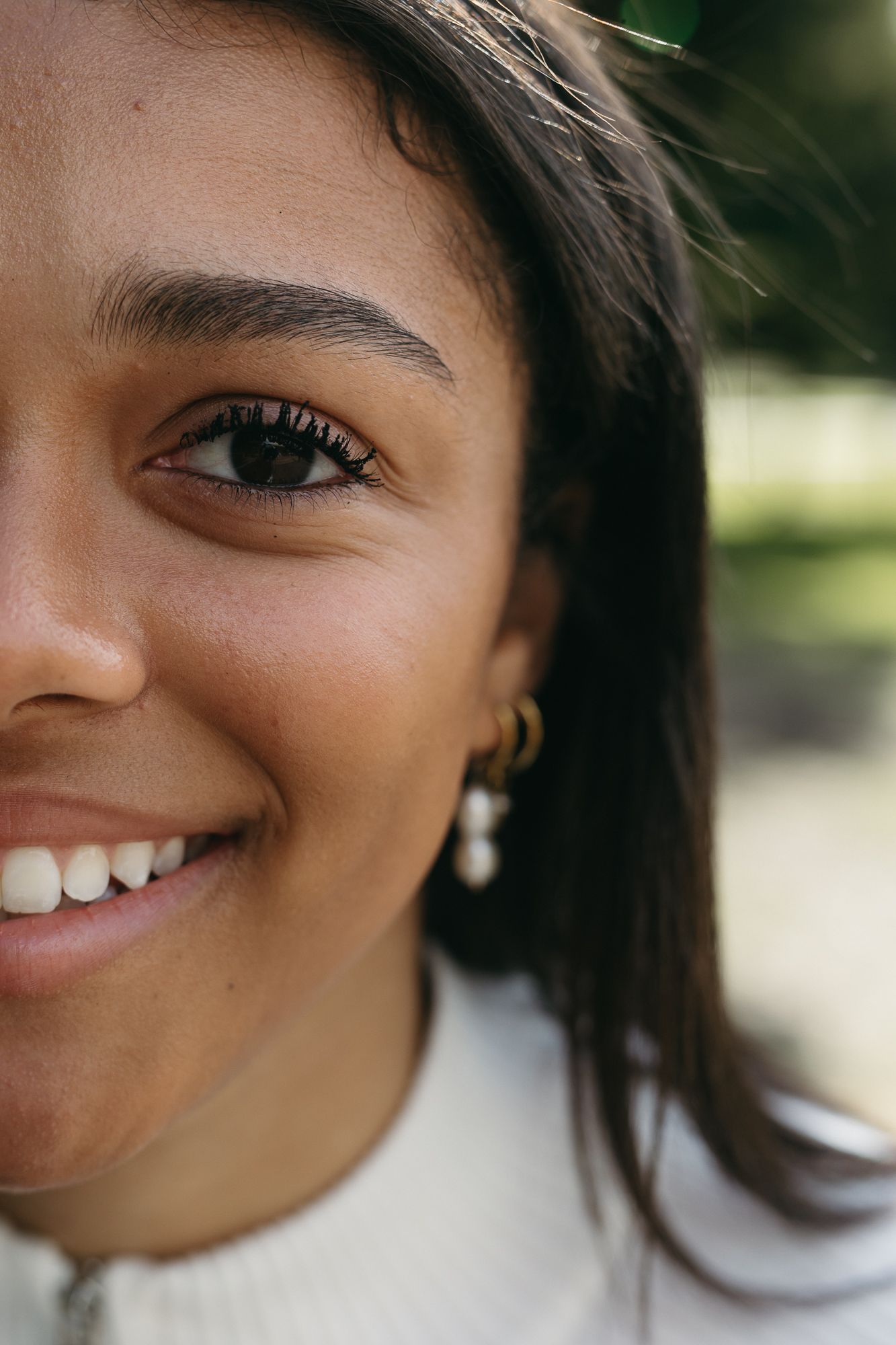 Close-up portrait of a smiling young woman with dark hair wearing gold hoop and pearl earrings outdoors.
