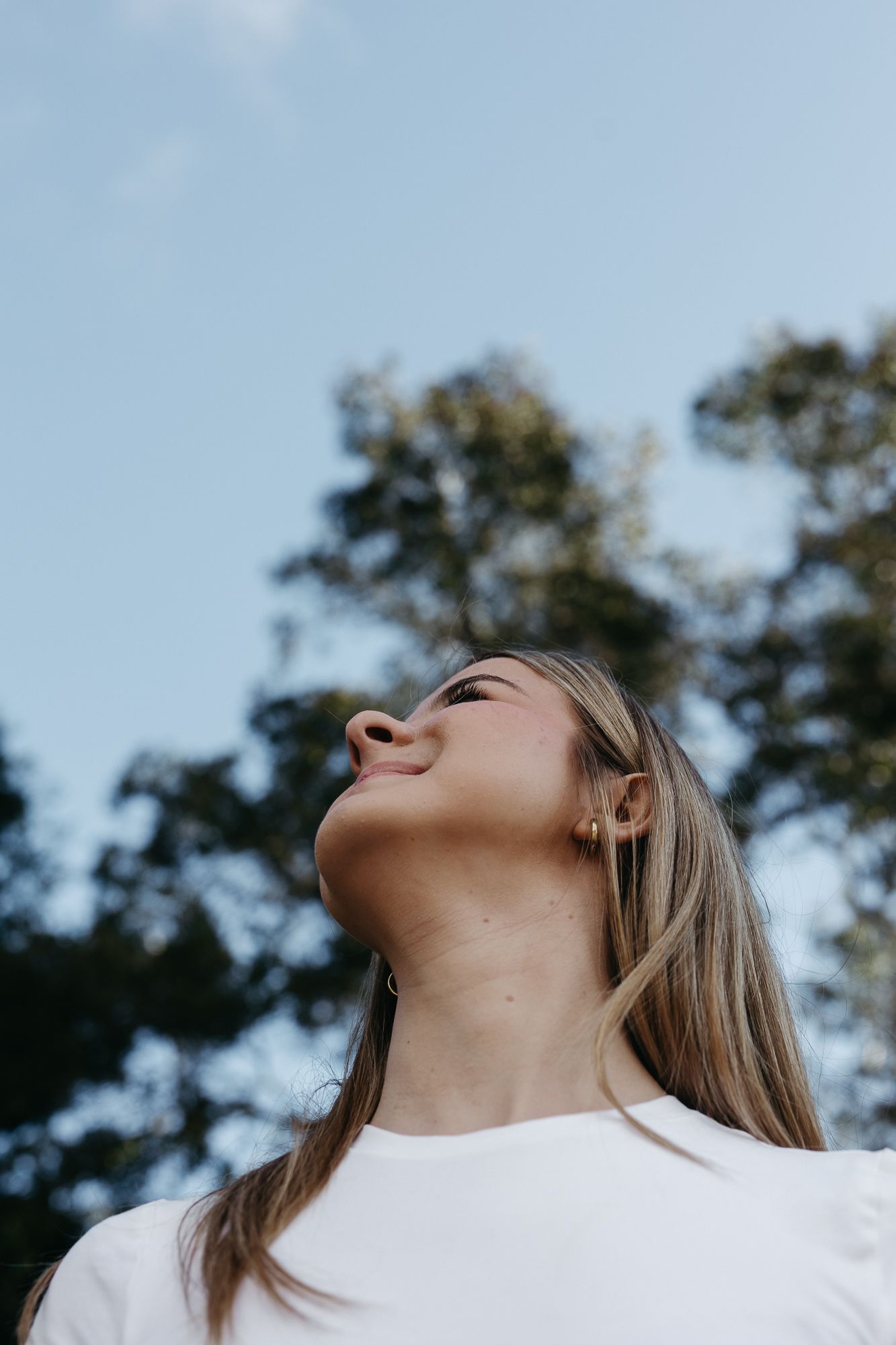 Young woman with long blonde hair smiling upward outdoors, wearing a white t-shirt with trees and blue sky behind her.