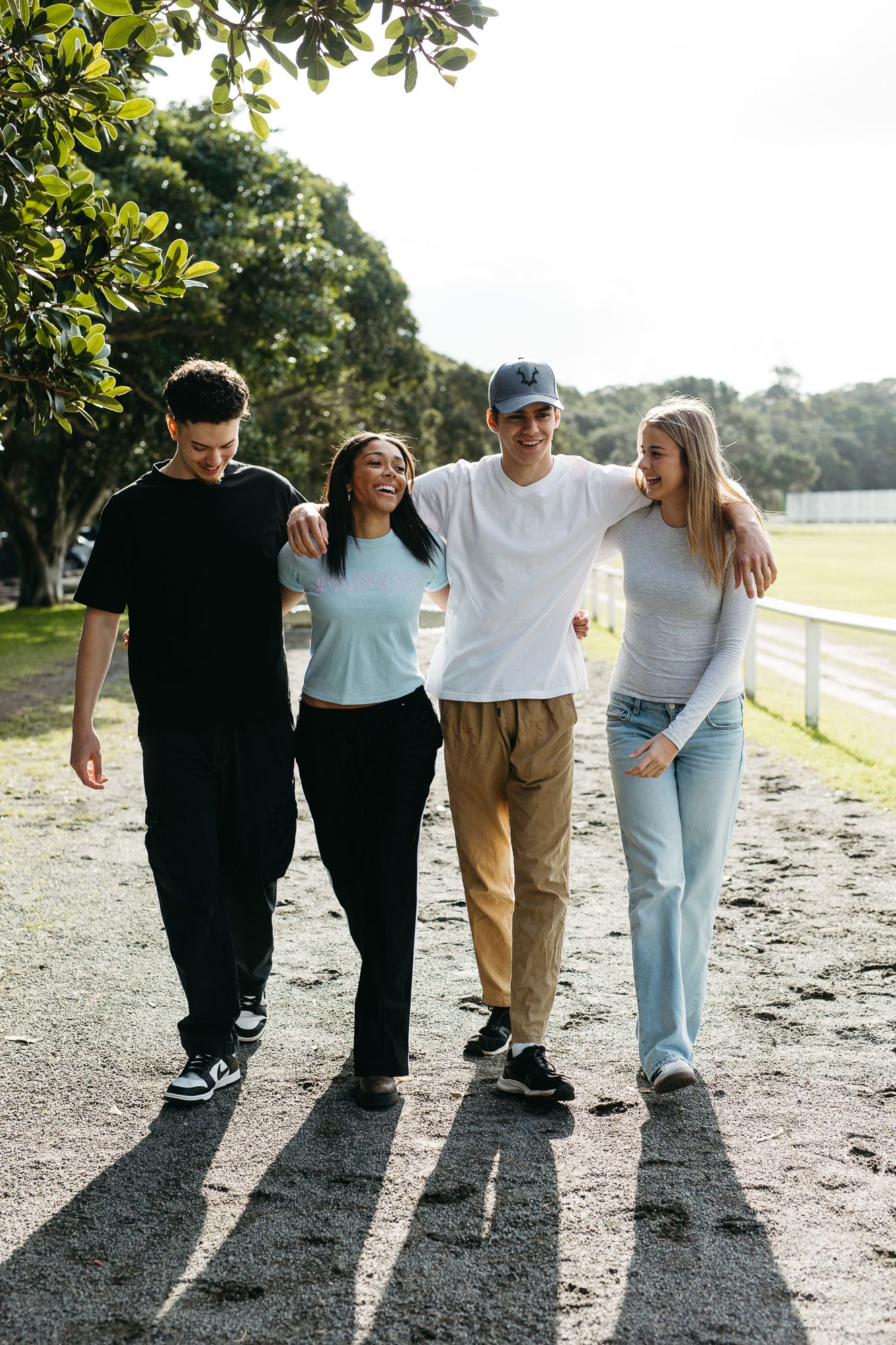 Four friends walking together outdoors, arms around each other, smiling and laughing on a sunny day.