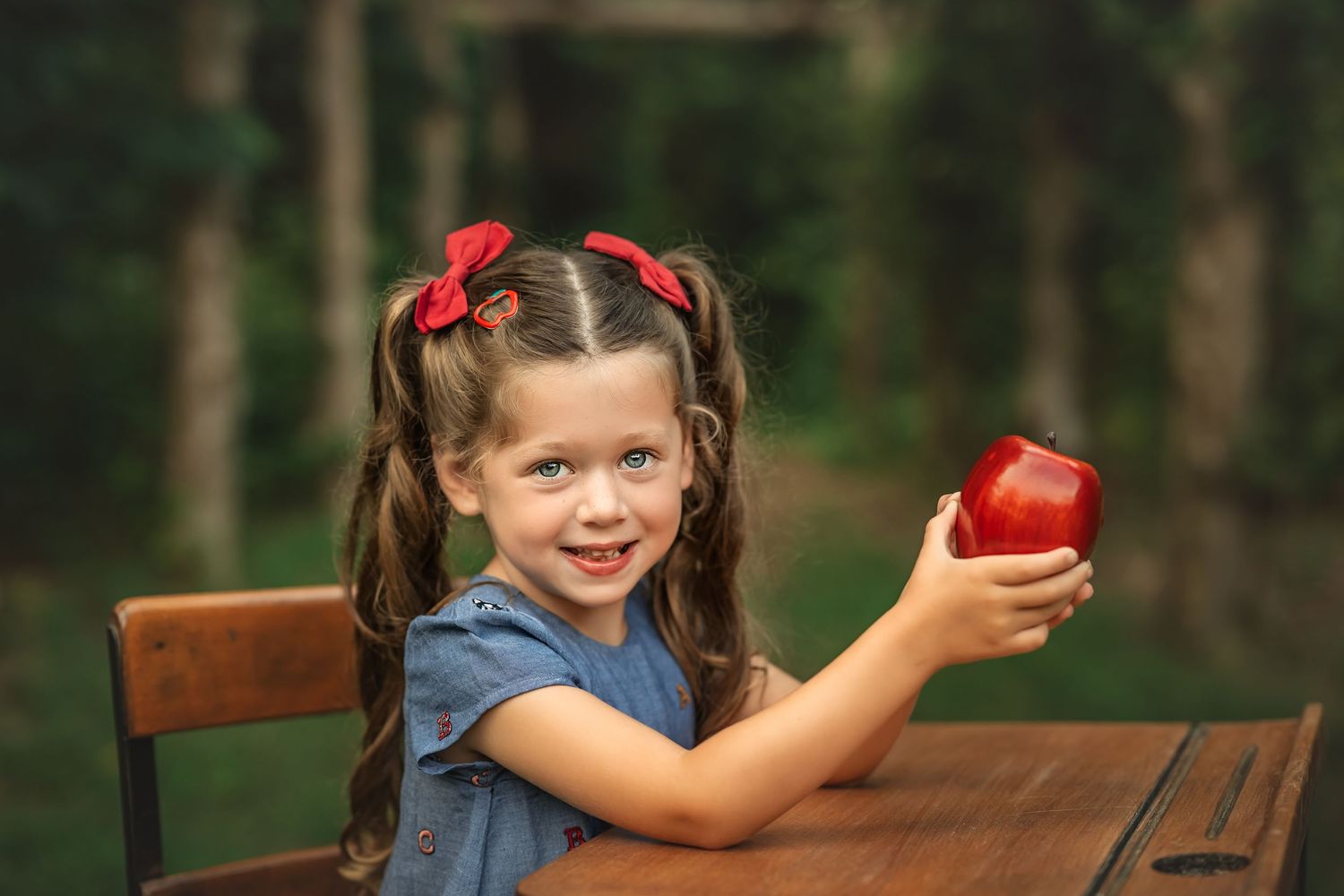 A smiling little girl with pigtails and red bows holds a red apple at a wooden school desk outdoors for back to school photos.