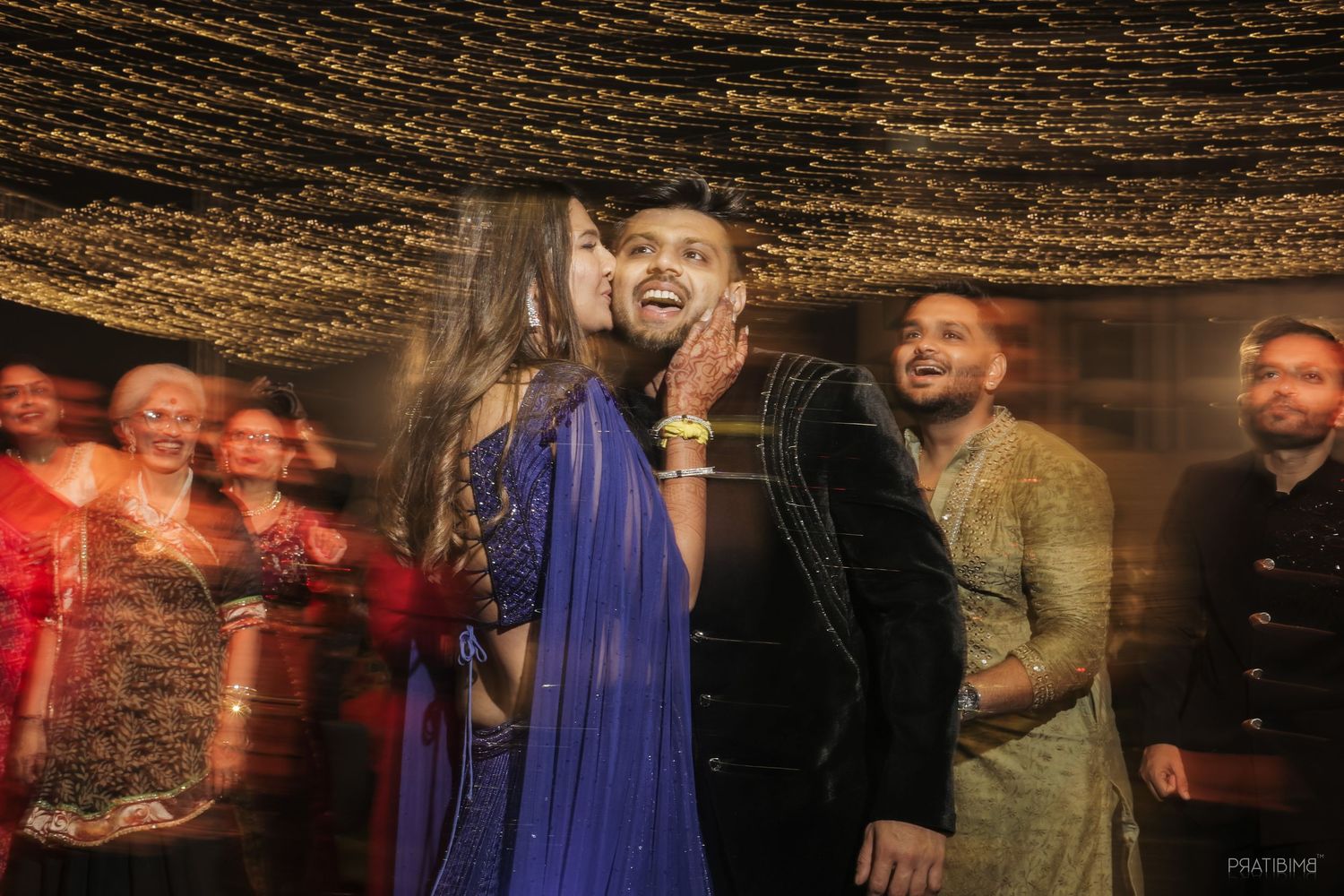 Couple sharing a romantic moment at an Indian wedding celebration with guests in colorful attire.