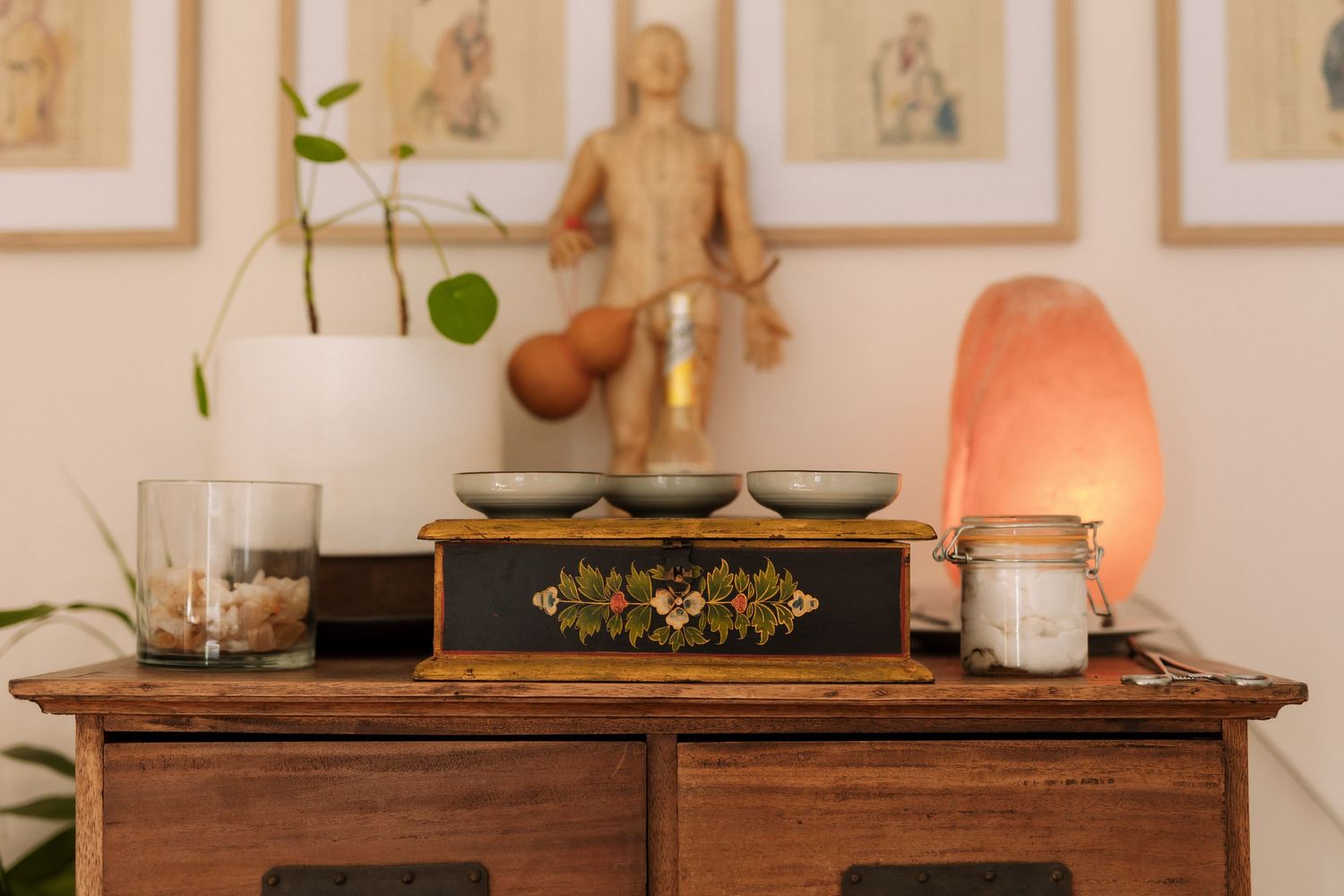 Wooden cabinet with Himalayan salt lamp, singing bowls, candles, and acupuncture chart in a serene wellness studio.