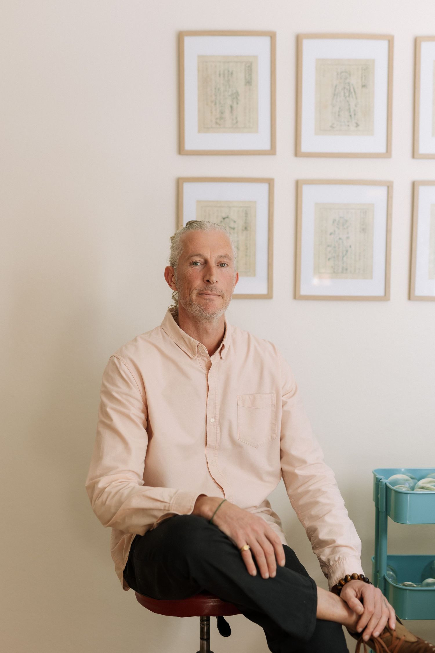 Middle-aged man with short gray hair sits on a stool in a light pink shirt, with framed artwork on the wall behind him.