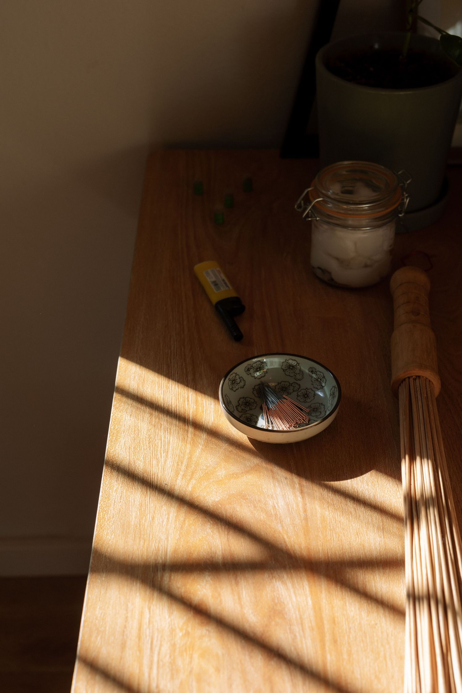 Wooden table with matcha bowl, rolling pin, glass jar, and dropper bottle in warm sunlight with diagonal shadows.