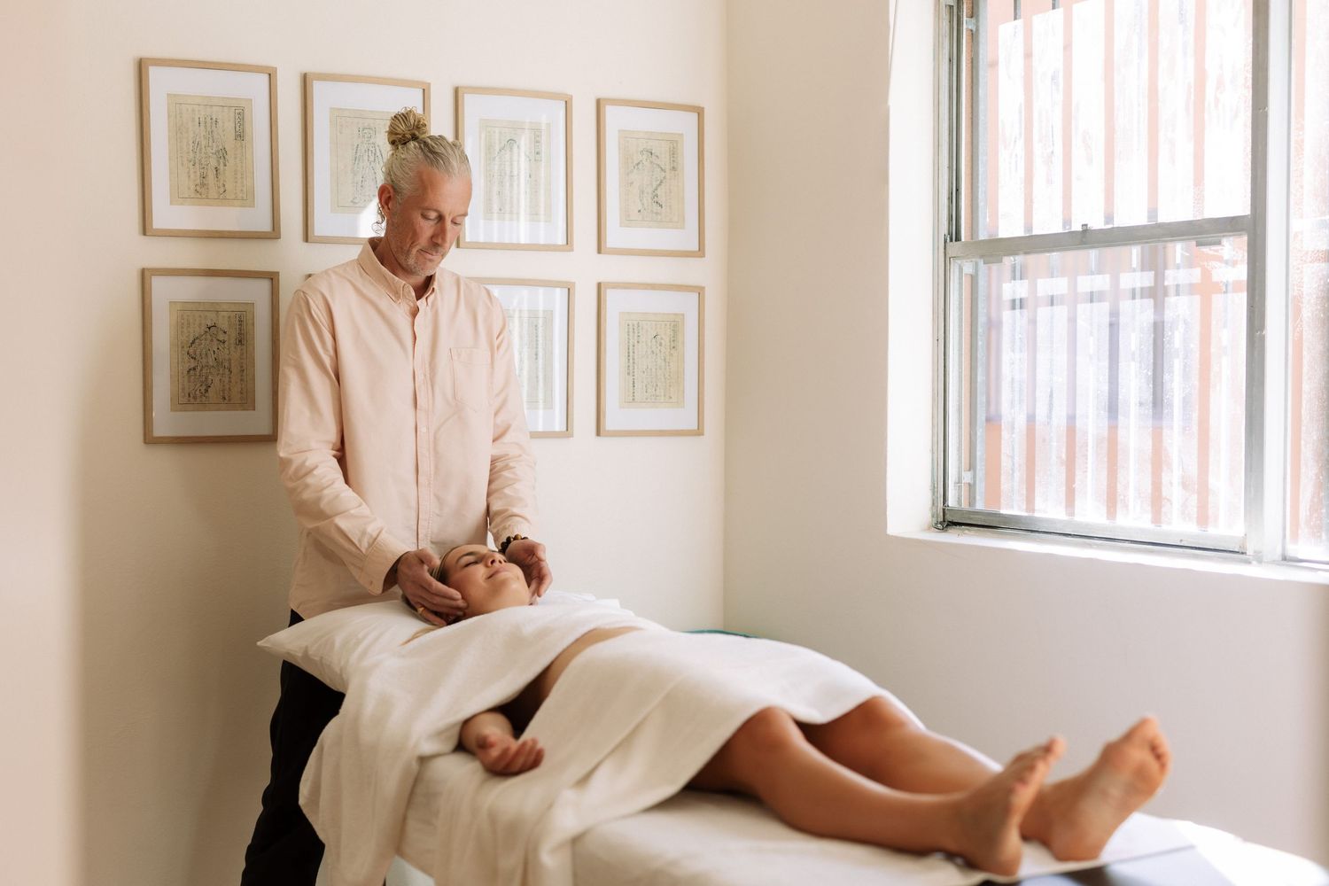 A massage therapist stands beside a client lying on a treatment table in a serene, light-filled spa room with framed art.