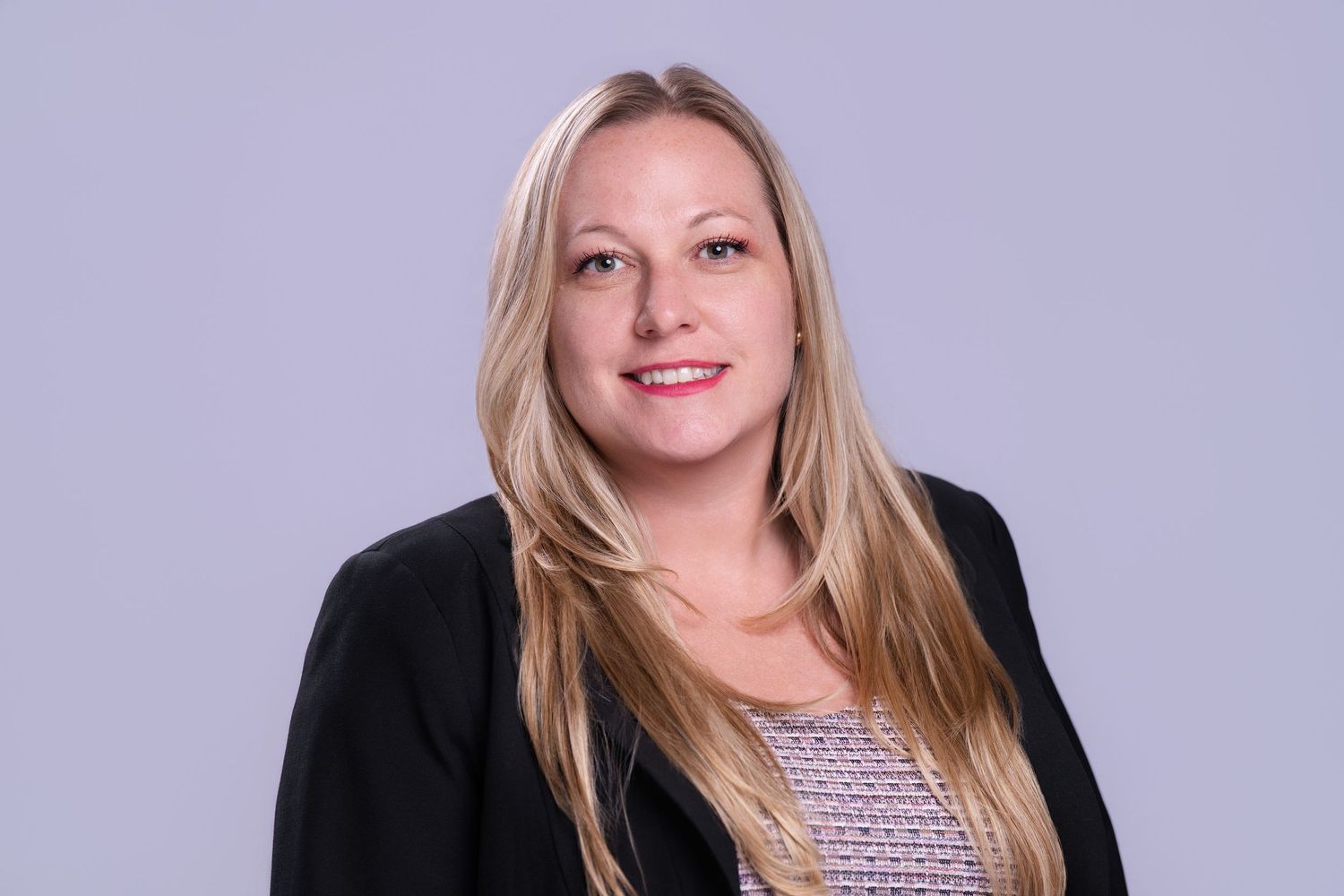 Professional headshot of a smiling blonde woman in a black blazer against a gray background.
