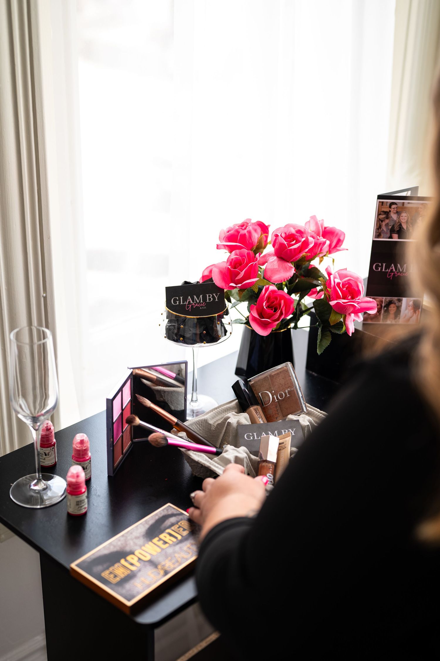 Glamorous vanity desk with pink roses, Dior makeup, brushes, and Glam By branding near a bright window.