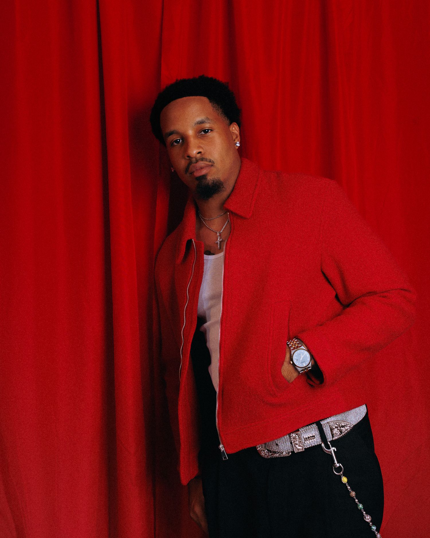 Young man in red jacket and black pants sits relaxed on black leather sofa against red curtain backdrop.