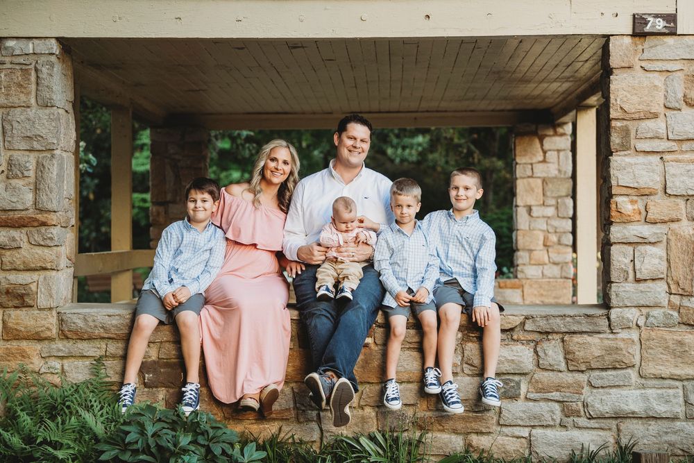 Family of six sitting under a stone pergola, parents with four boys in coordinated blue and pink outfits.