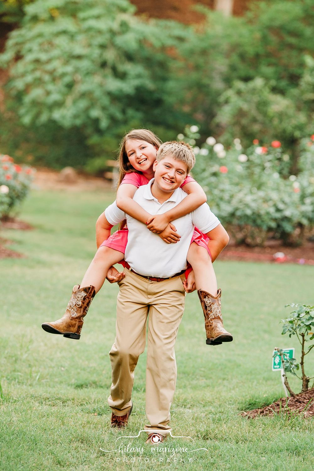 Boy giving girl a piggyback ride in a garden, both smiling, girl wearing cowboy boots, lush green background.