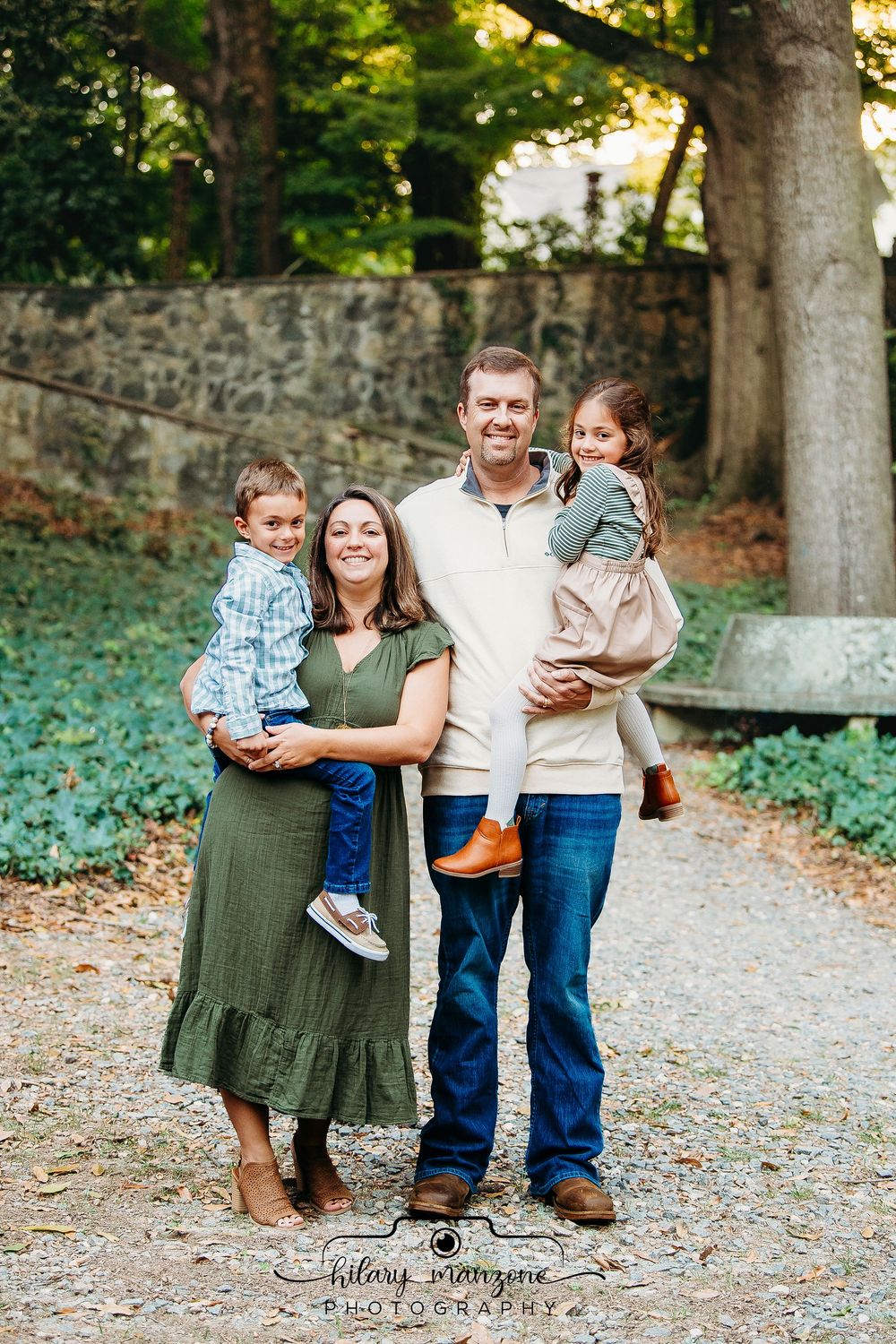 Family of four smiling outdoors, parents holding two young children, posing on a gravel path surrounded by greenery.