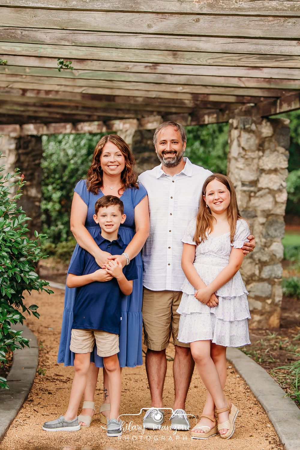 A smiling family of four poses outdoors under a wooden pergola, wearing coordinating blue and neutral outfits.