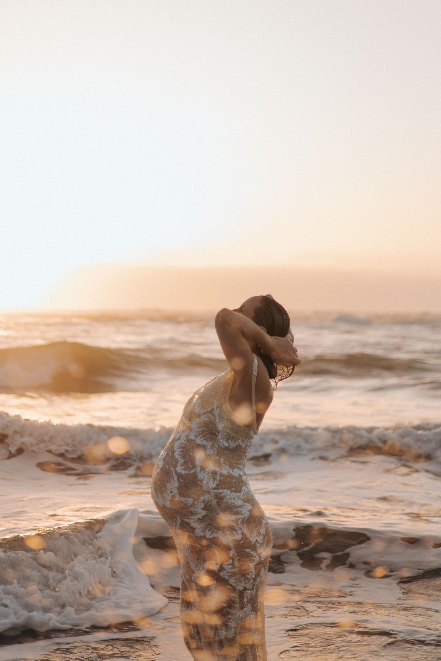 Woman in floral dress standing in ocean waves at golden sunset, hands raised to head, warm light glowing.