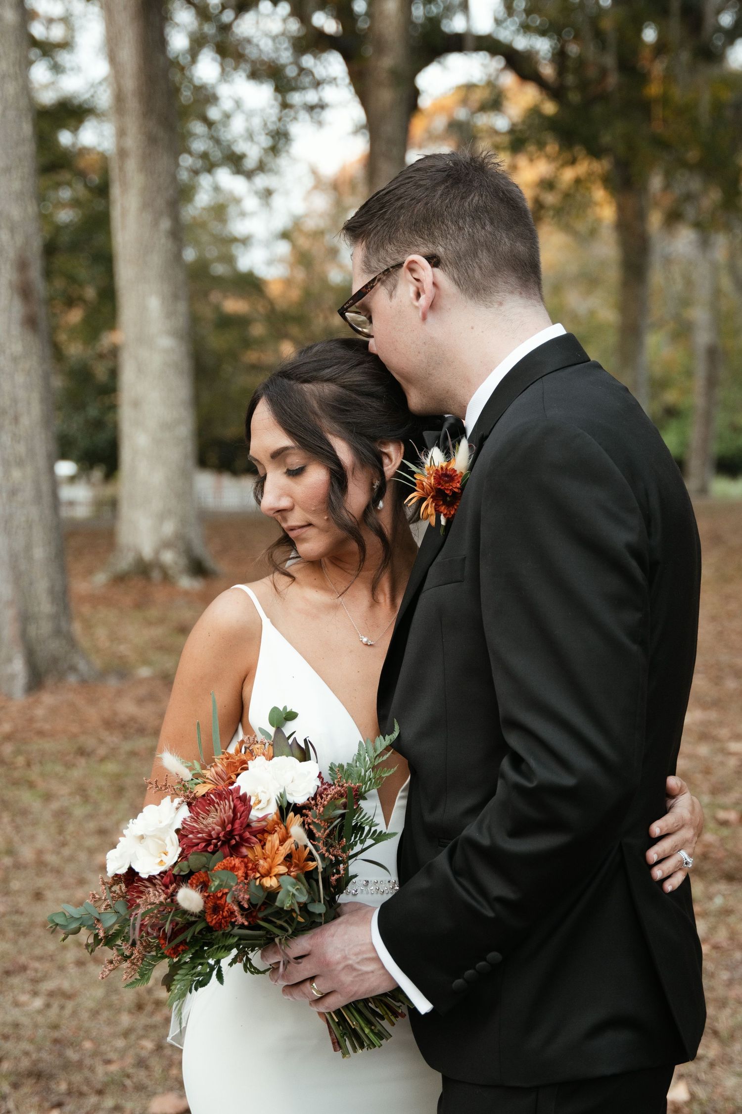 Bride and groom embrace outdoors among autumn trees, bride holding a rich burgundy and white fall wedding bouquet.