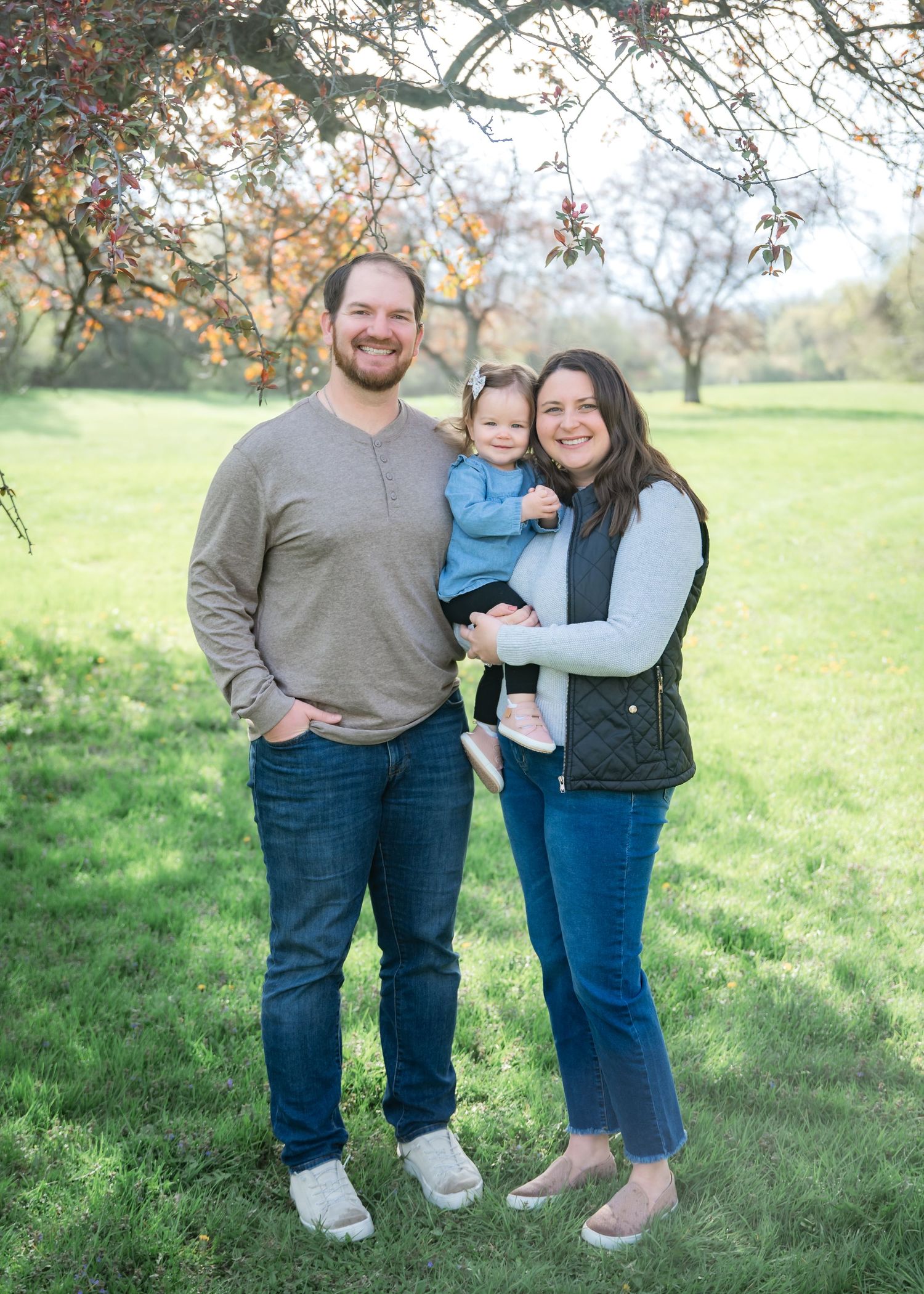 A smiling family of three poses outdoors in a green park, with a man, woman holding a toddler, and autumn trees behind them.
