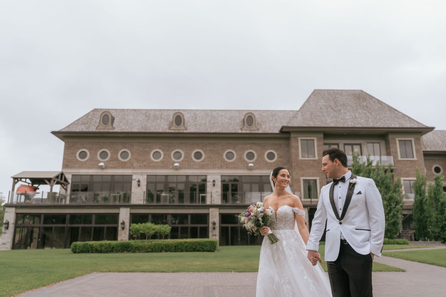 Groom carries bride in wedding dress on urban street with red bus and brick hotel building in background.