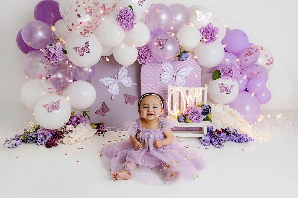 Baby girl in purple dress sits among lavender and white balloon arch with butterfly decorations for first birthday.