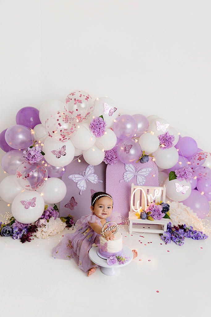 Smiling baby in purple tulle dress poses with first birthday cake under butterfly balloon garland backdrop.