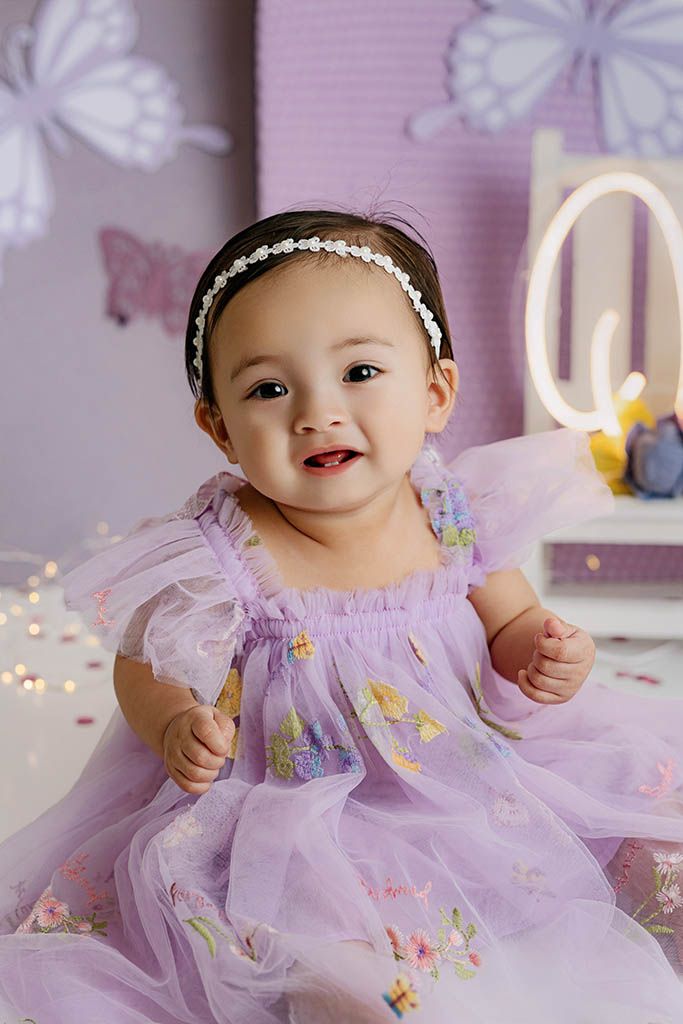 A baby girl in a lavender tulle dress with floral embroidery and pearl headband sits smiling at her first birthday photoshoot.