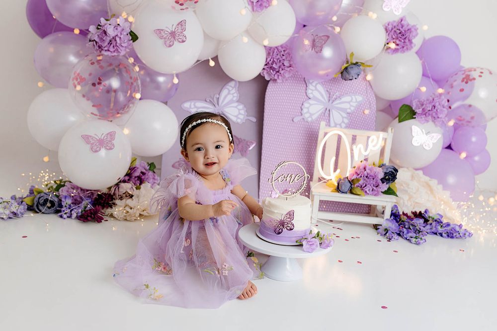 Baby girl in purple tutu dress sits beside a butterfly-themed first birthday cake with lavender balloon arch backdrop.