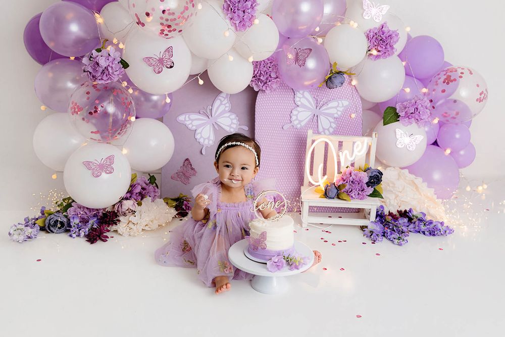 Baby girl in purple dress smiles at first birthday cake smash with butterfly balloon arch backdrop.