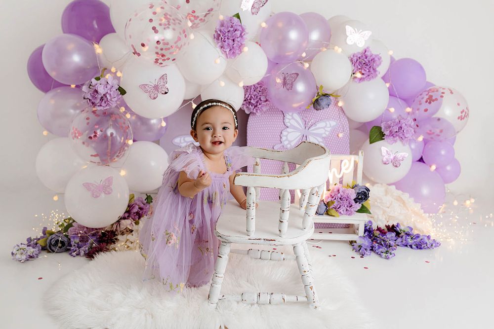 Toddler girl in purple tulle dress stands by white chair with butterfly balloon arch backdrop.
