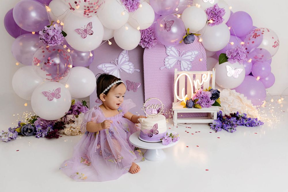 Toddler in lavender tulle dress reaches for butterfly birthday cake at purple balloon decorated party.