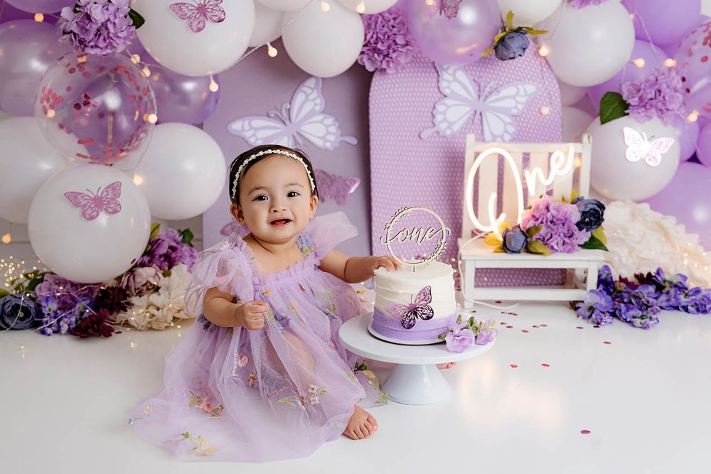 Happy baby in purple dress sits beside butterfly cake at first birthday smash with balloon arch.