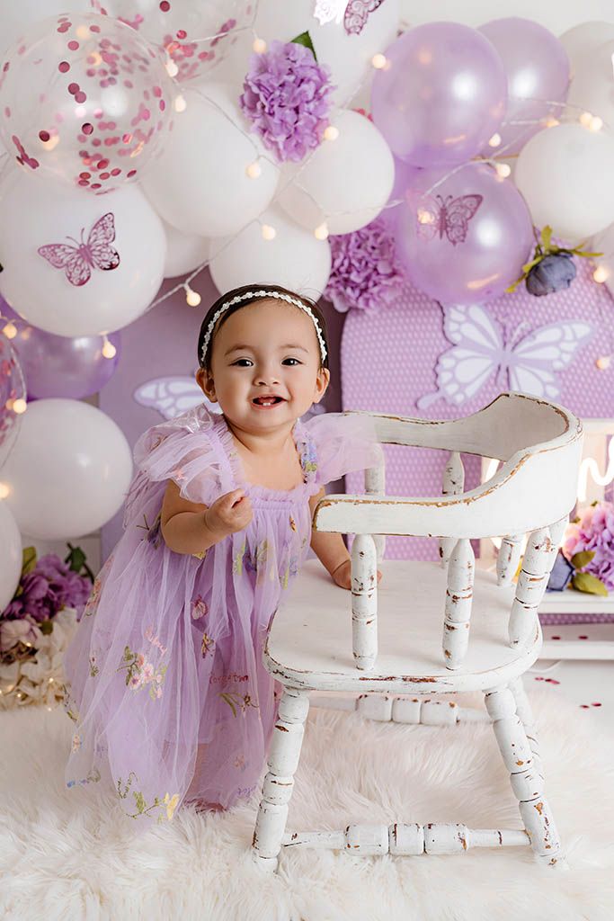 Smiling toddler in purple tulle dress stands beside rustic white chair with balloon backdrop.