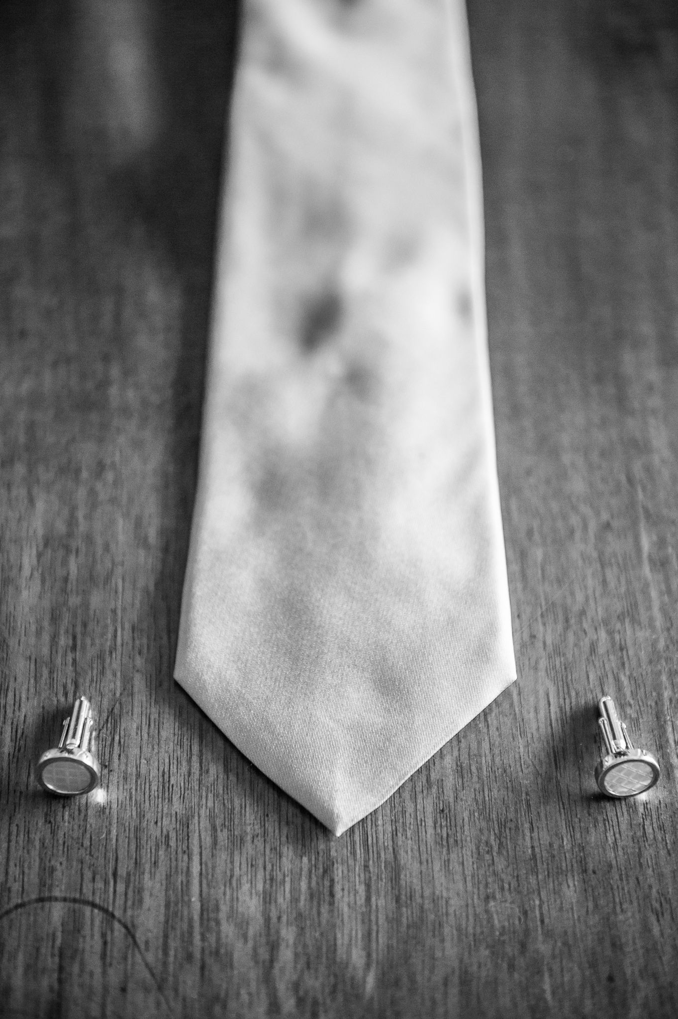 Black and white close-up of a silk wedding tie and silver cufflinks on a wooden surface.